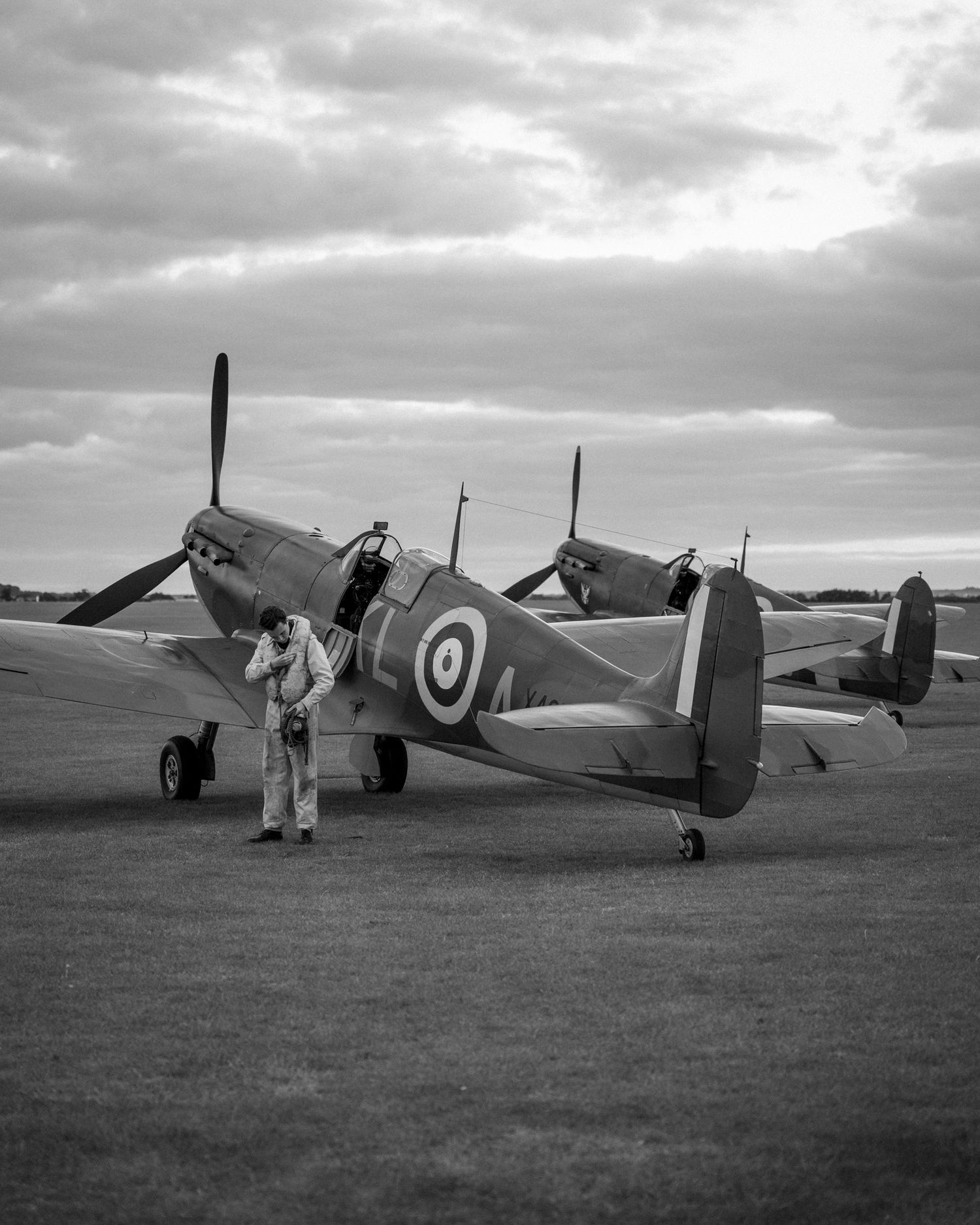 A vintage RAF Spitfire aircraft sits on grass airfield under cloudy skies.
