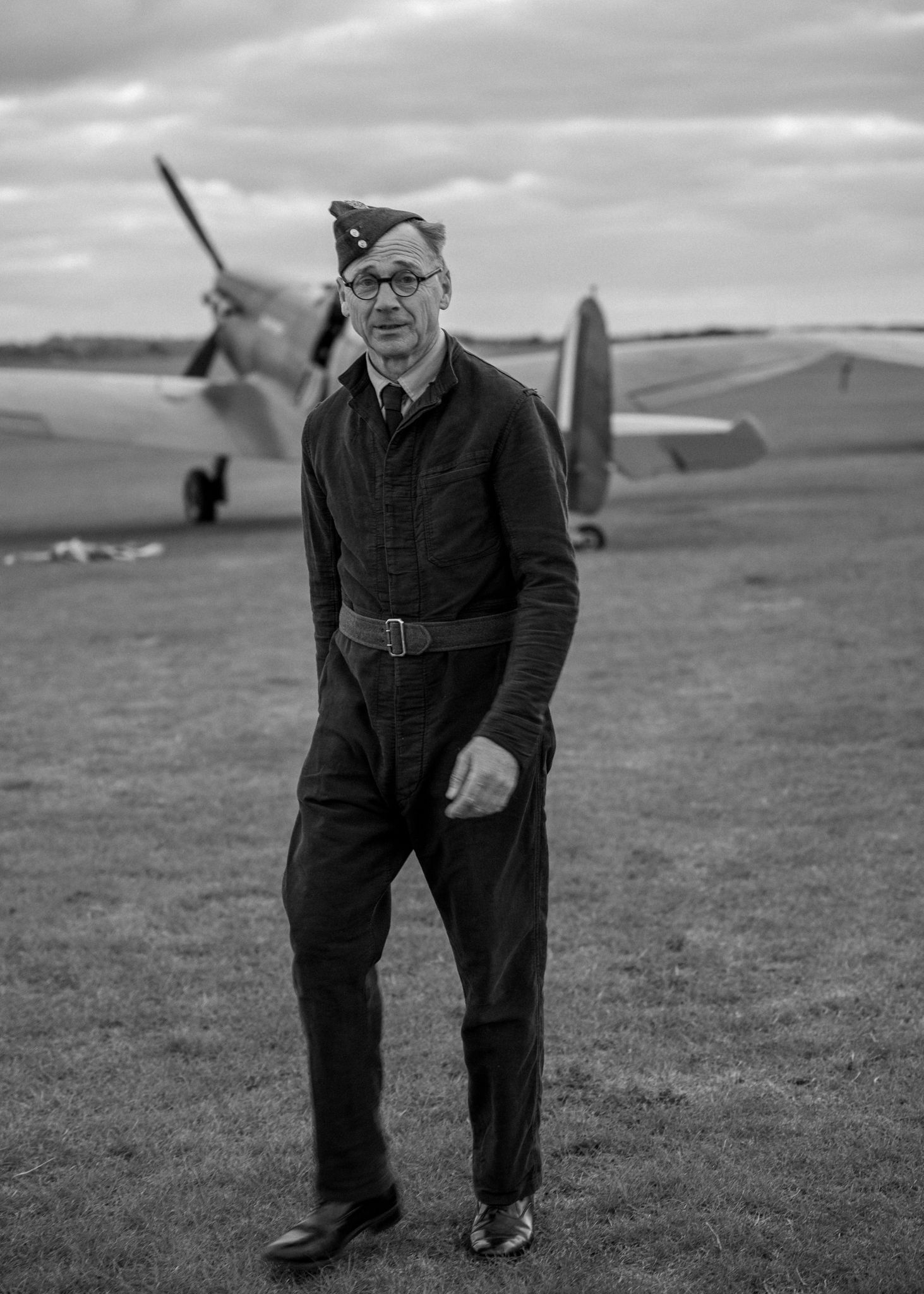 A person in vintage aviation attire stands in front of a classic Spitfire aircraft.
