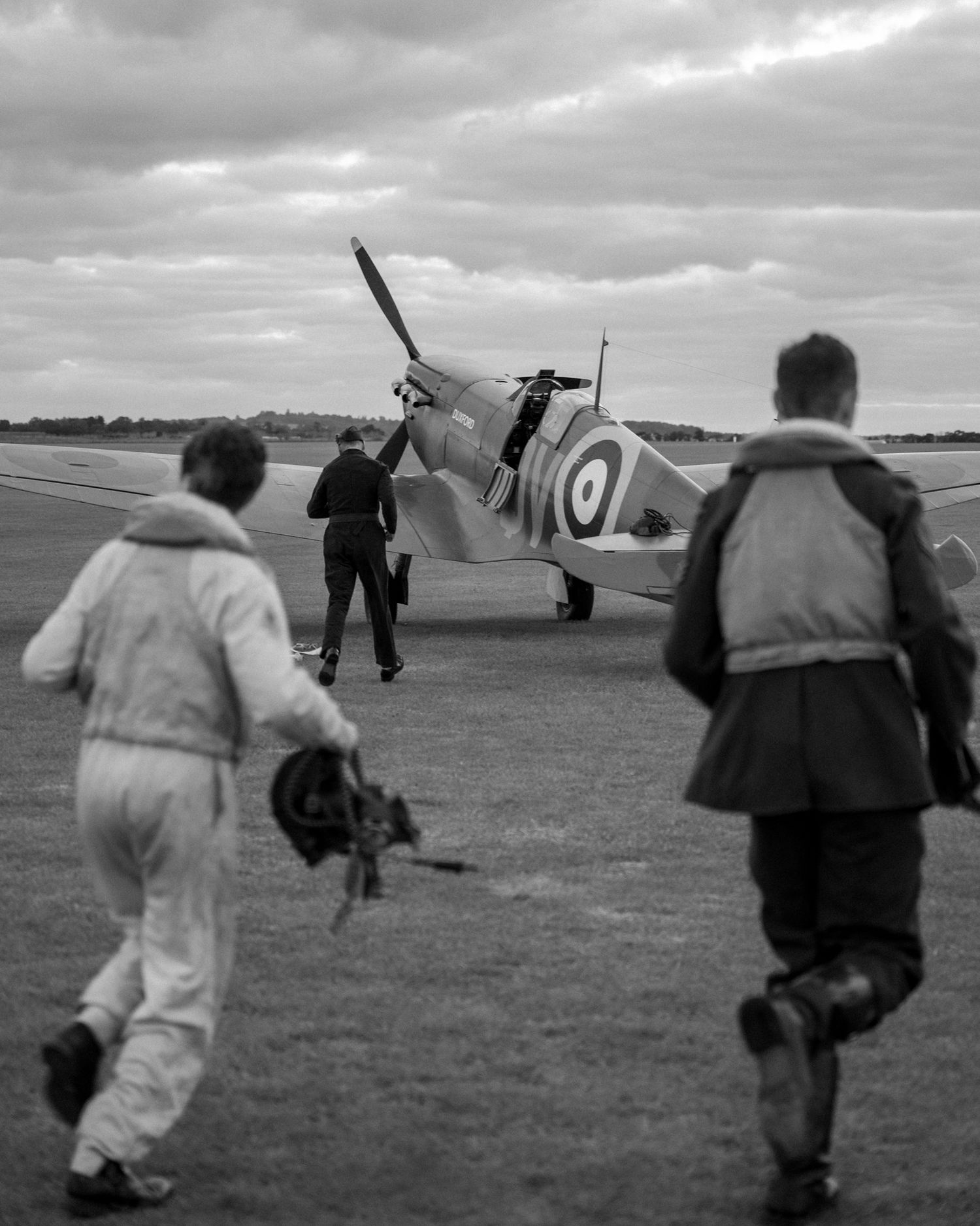 Three people approach a parked Spitfire aircraft on a grass airfield.
