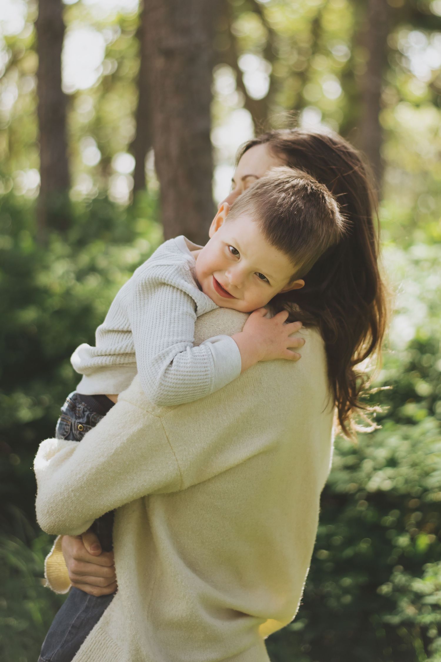 A mother and child share a tender embrace while standing in a sunlit forest with green foliage in the background.