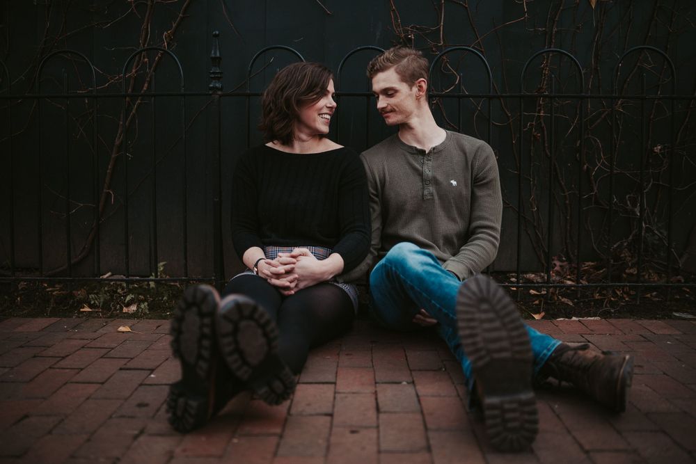 A couple in casual attire sits together on brick pavement against a dark wall during an intimate moment.