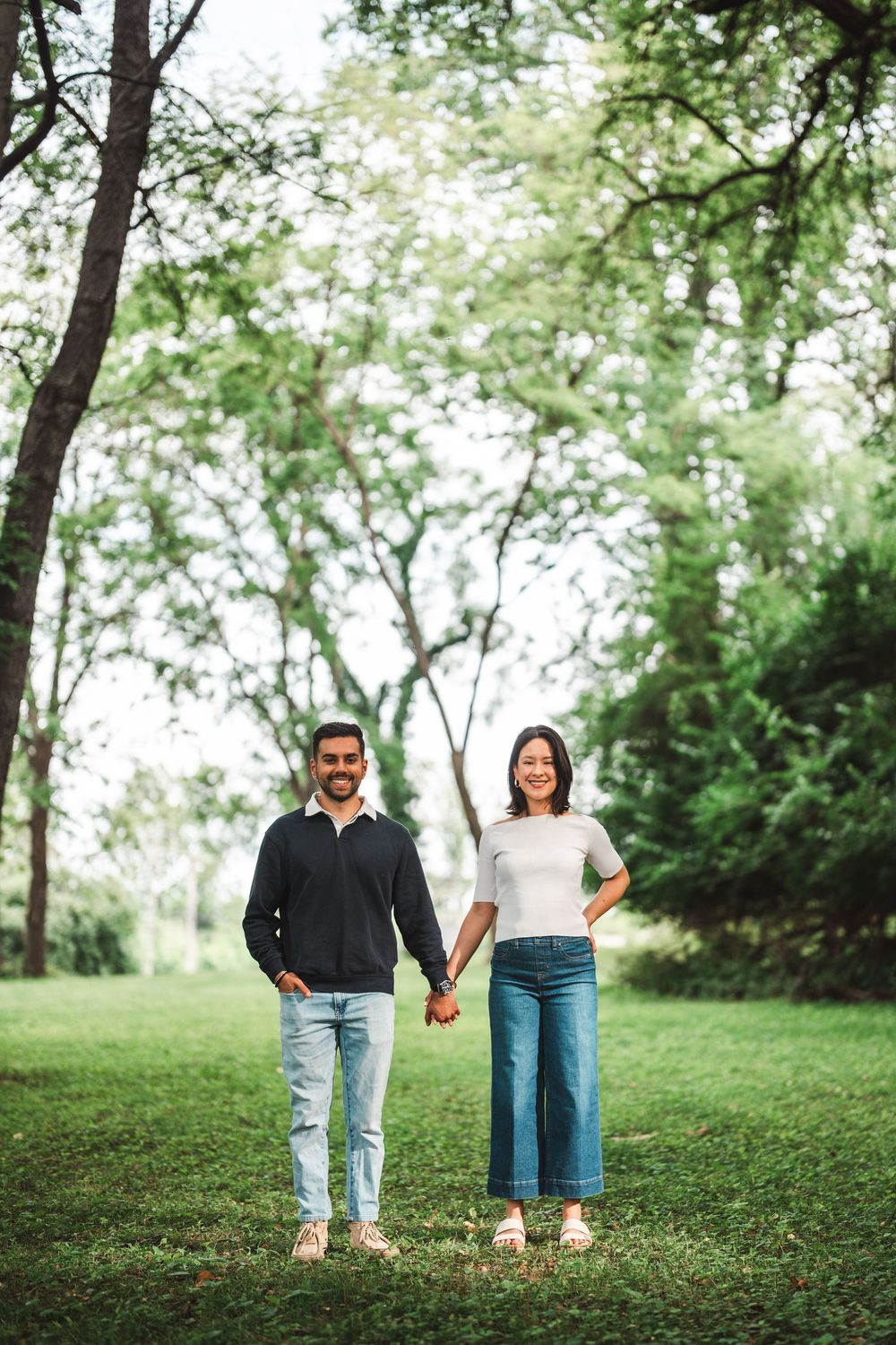 A couple holding hands stands on green grass in a park with tall trees creating a natural backdrop.