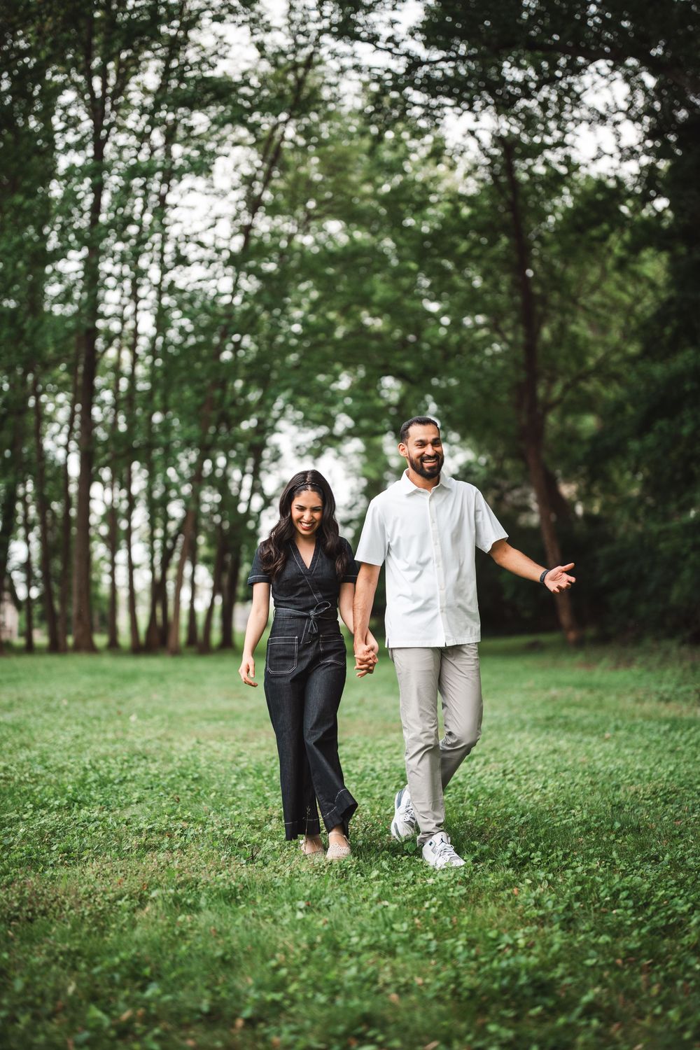 A couple walks together holding hands on lush green grass surrounded by tall trees in a park setting.