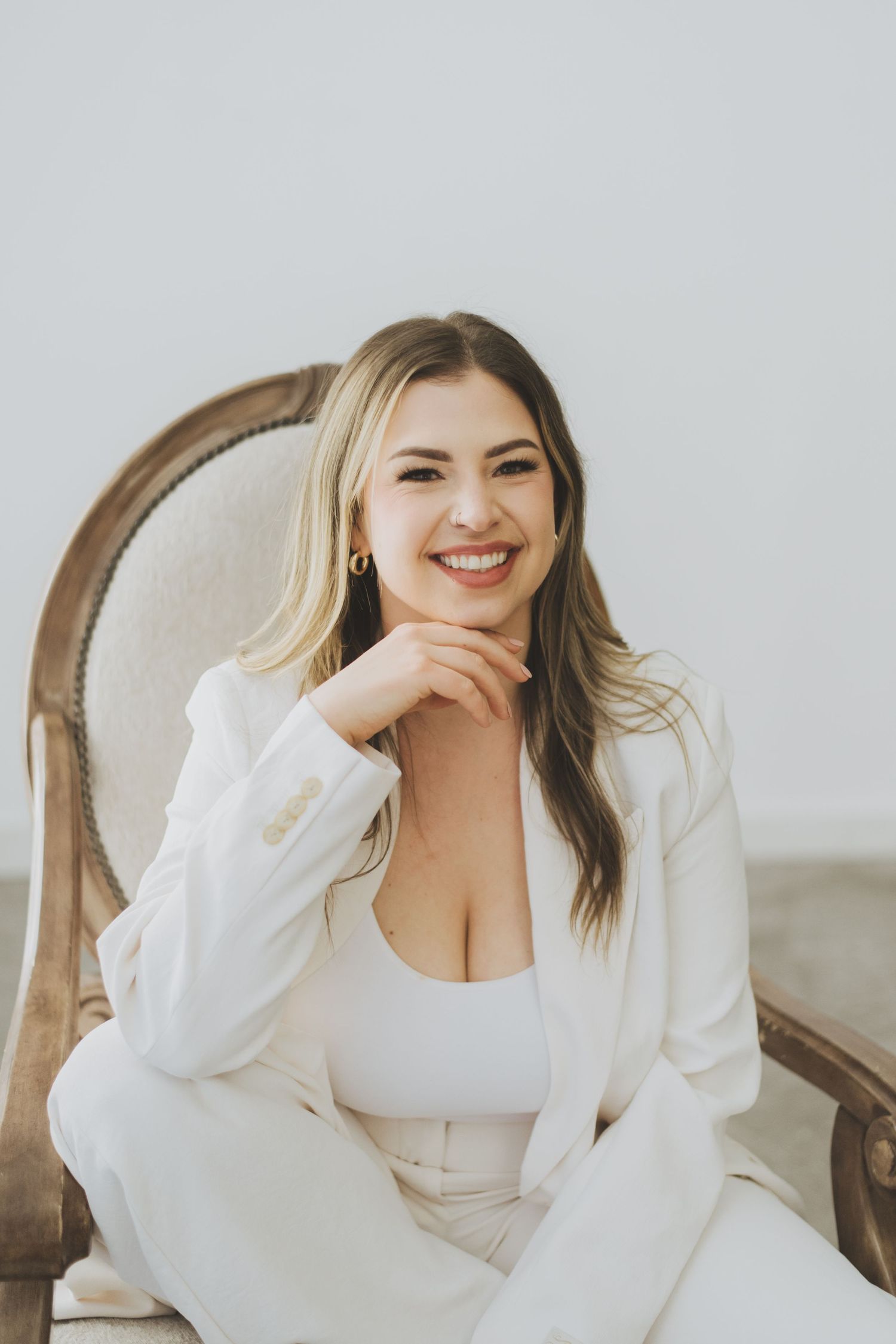 A woman in an all-white outfit sits in an antique chair, wearing a blazer over a fitted top with a relaxed pose.