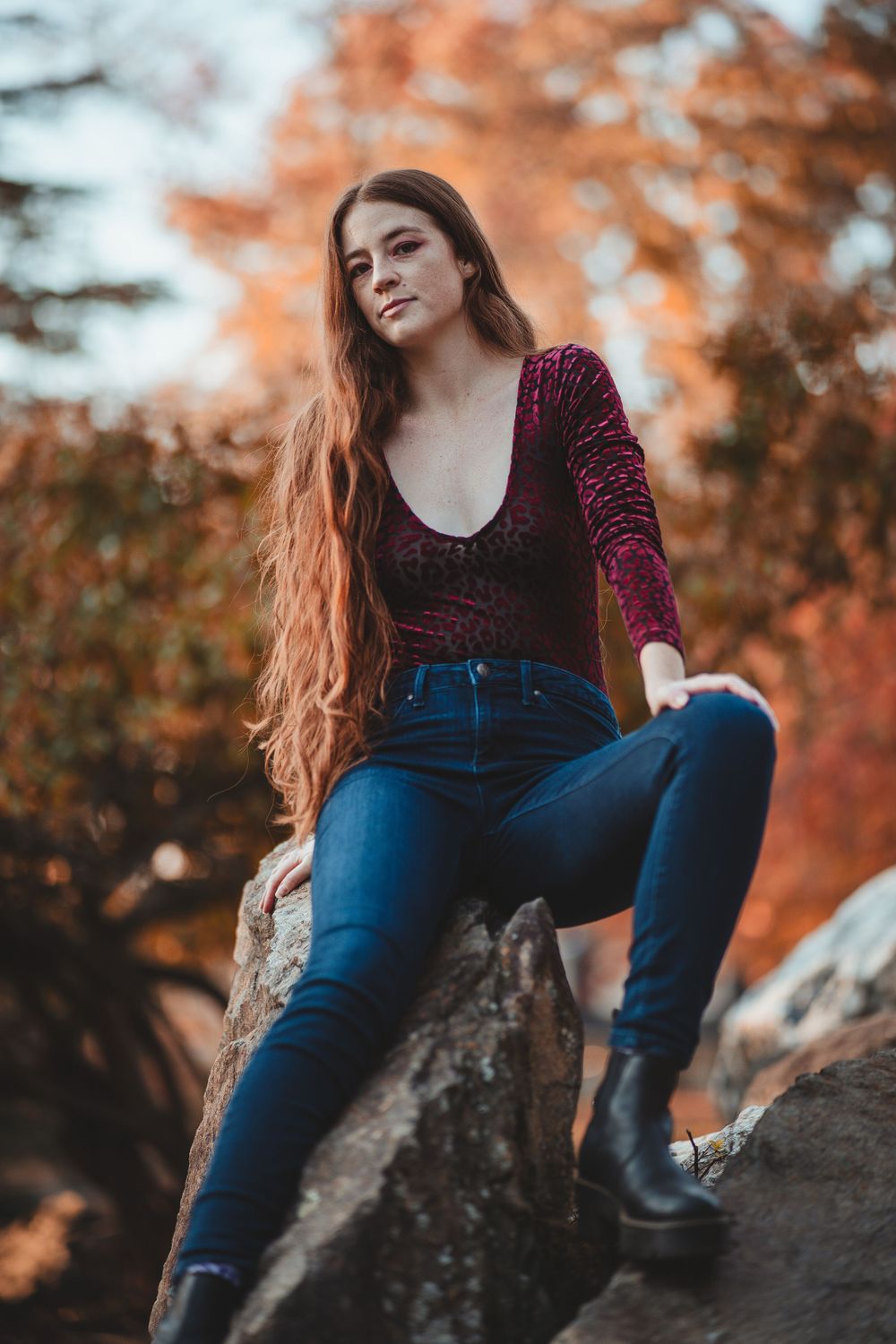 A woman in a burgundy velvet top and blue jeans sits on a rocky outcrop during autumn with orange foliage in background in North Bethesda.