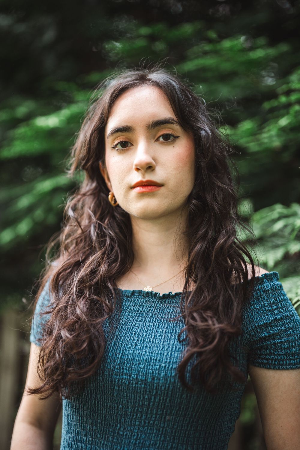 A portrait in a natural setting featuring long wavy dark hair, red lipstick and a blue smocked top against green foliage.