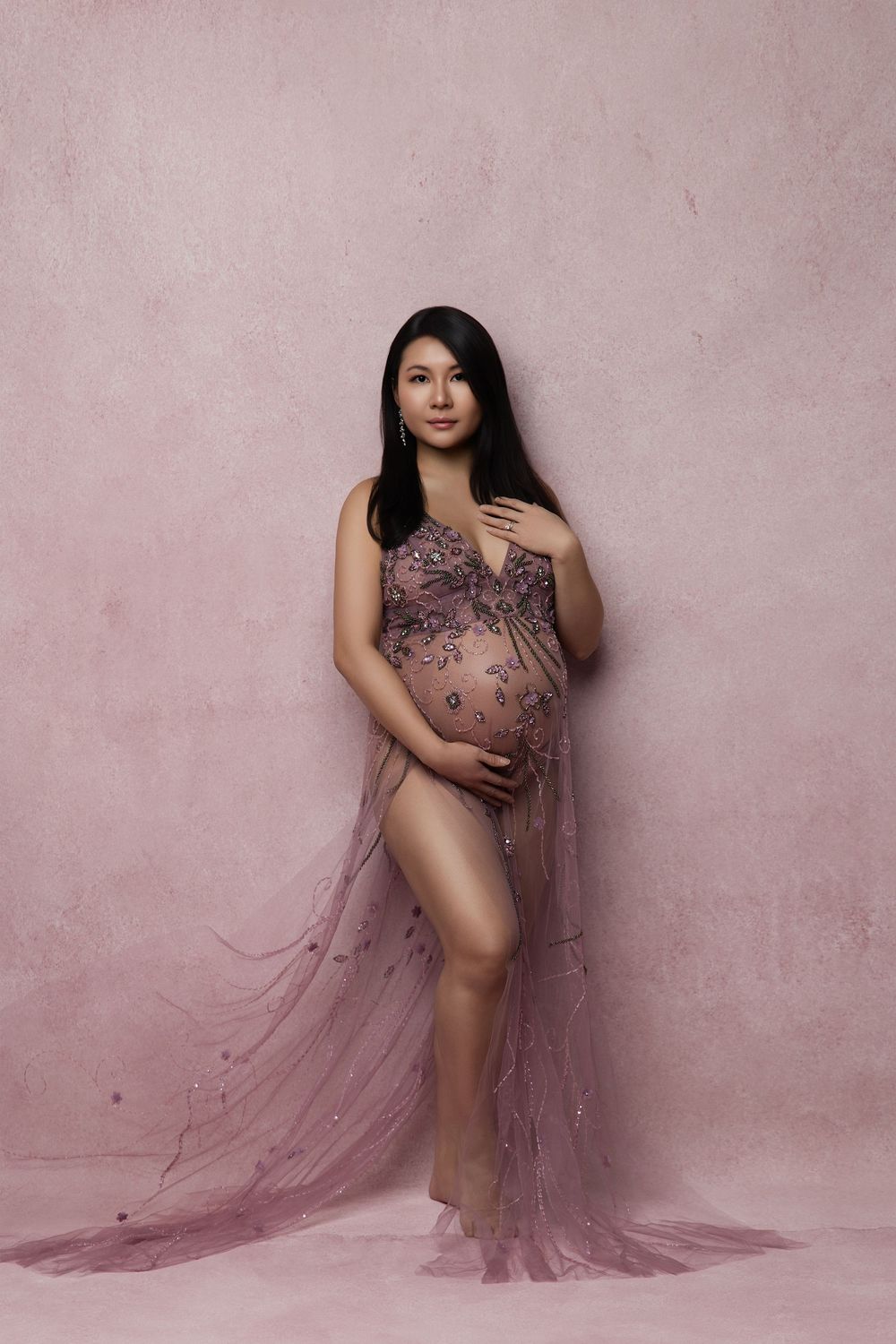 A pregnant woman poses in a sheer mauve maternity gown against a pink textured backdrop for an elegant portrait.