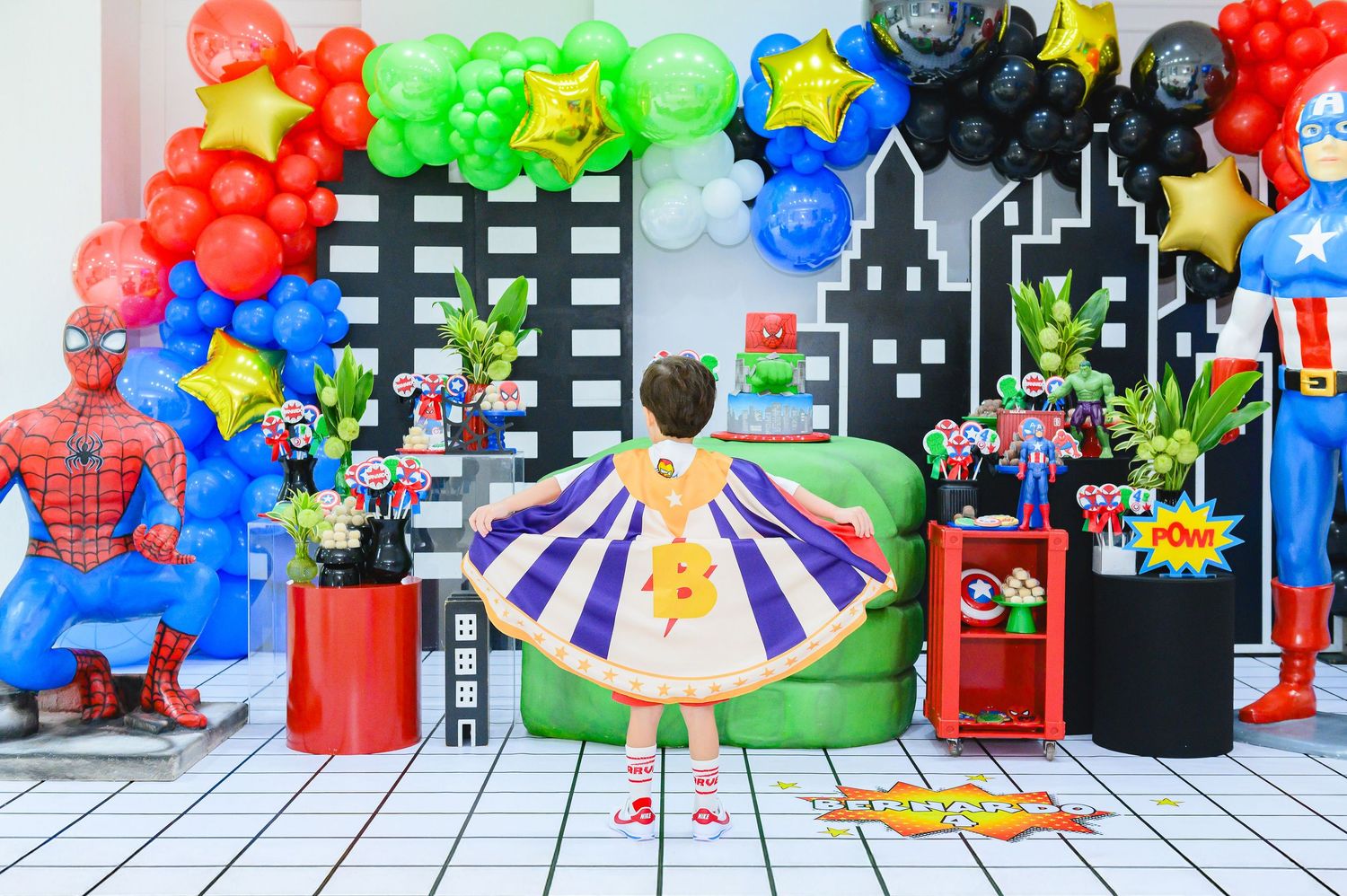 A superhero-themed birthday party setup with cityscape backdrop, colorful balloon arch, and festive decorations.