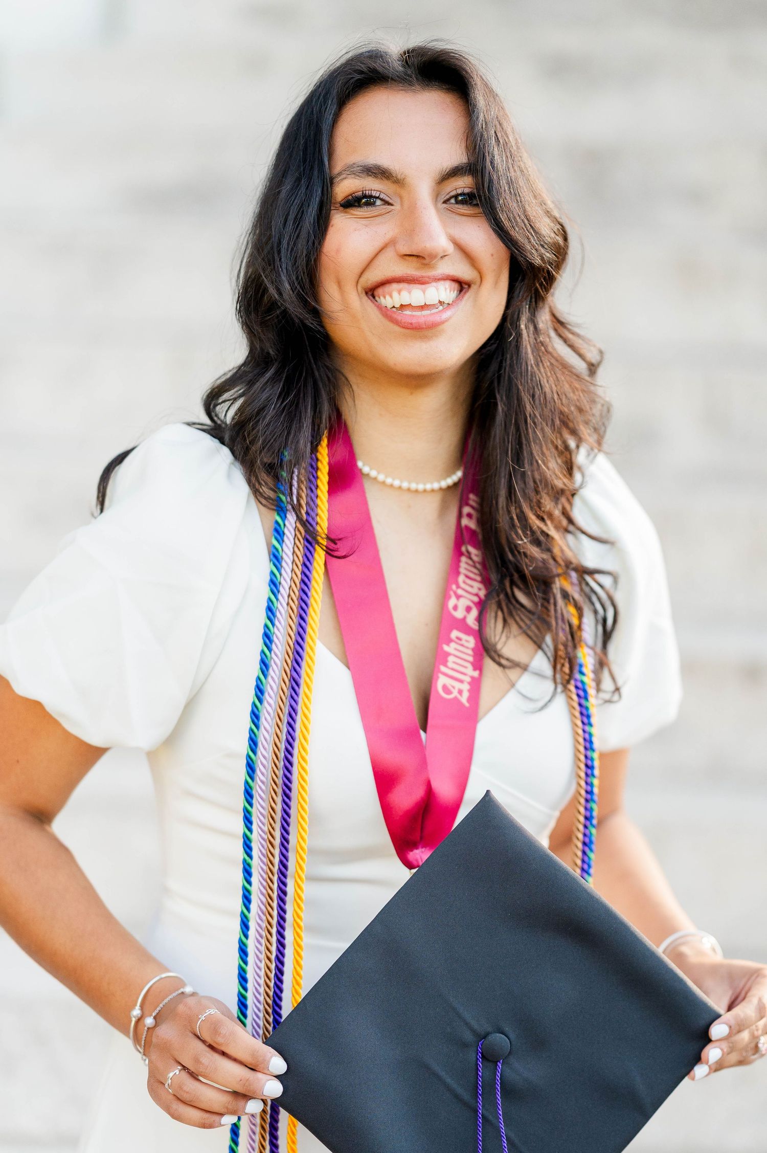 Graduate wearing white top and honor cords smiles while holding black graduation cap against light background.