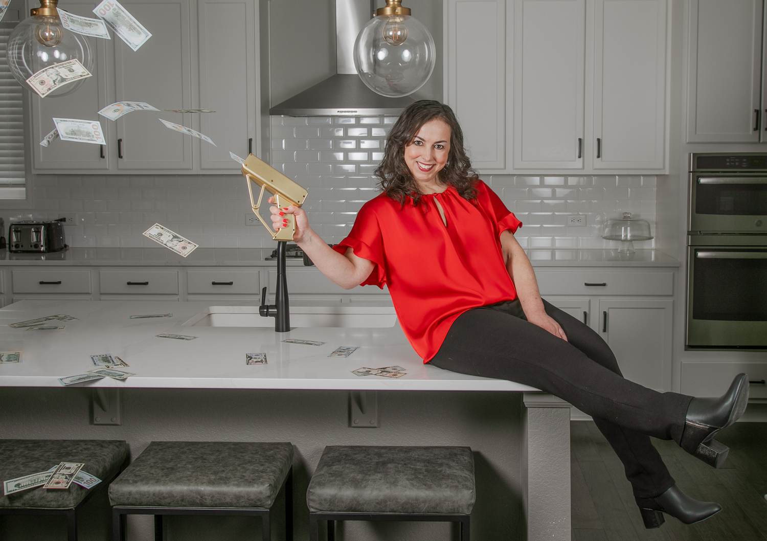 Someone in a red top sits casually on a kitchen counter while money bills float through the air in a modern grey kitchen.