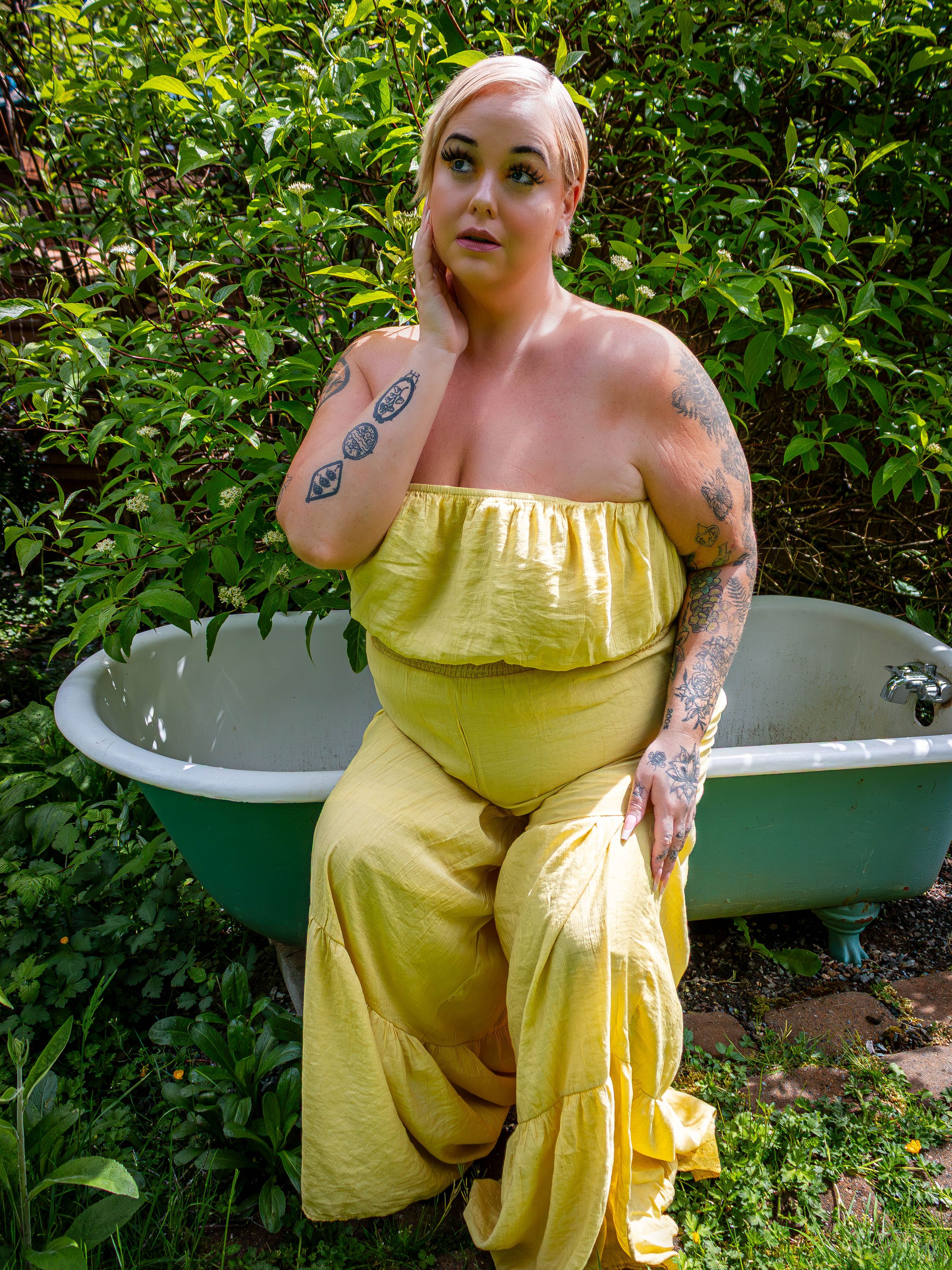 A person in a yellow strapless jumpsuit poses against a green garden backdrop with a vintage bathtub.