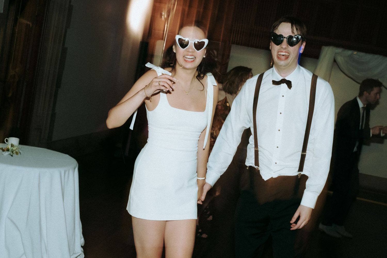 Stylish couple in wedding attire wearing sunglasses pose with drinks at reception.