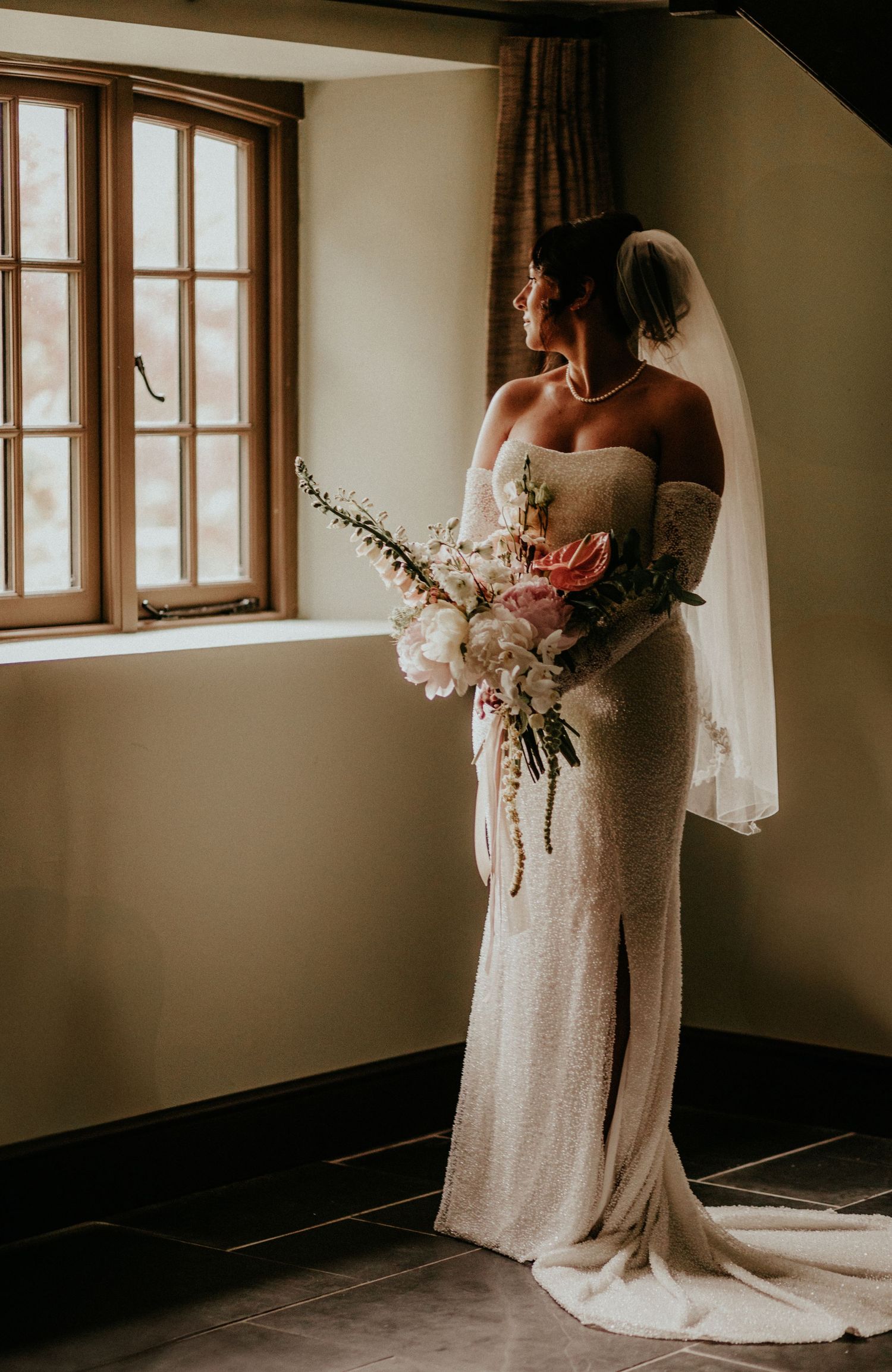 A bride in a flowing white dress holds a romantic bouquet of flowers while standing near a window in soft natural light.