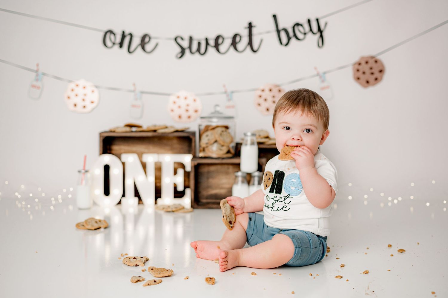 First birthday photo setup with white marquee letters, cookie decorations, and wooden crates against white backdrop.
