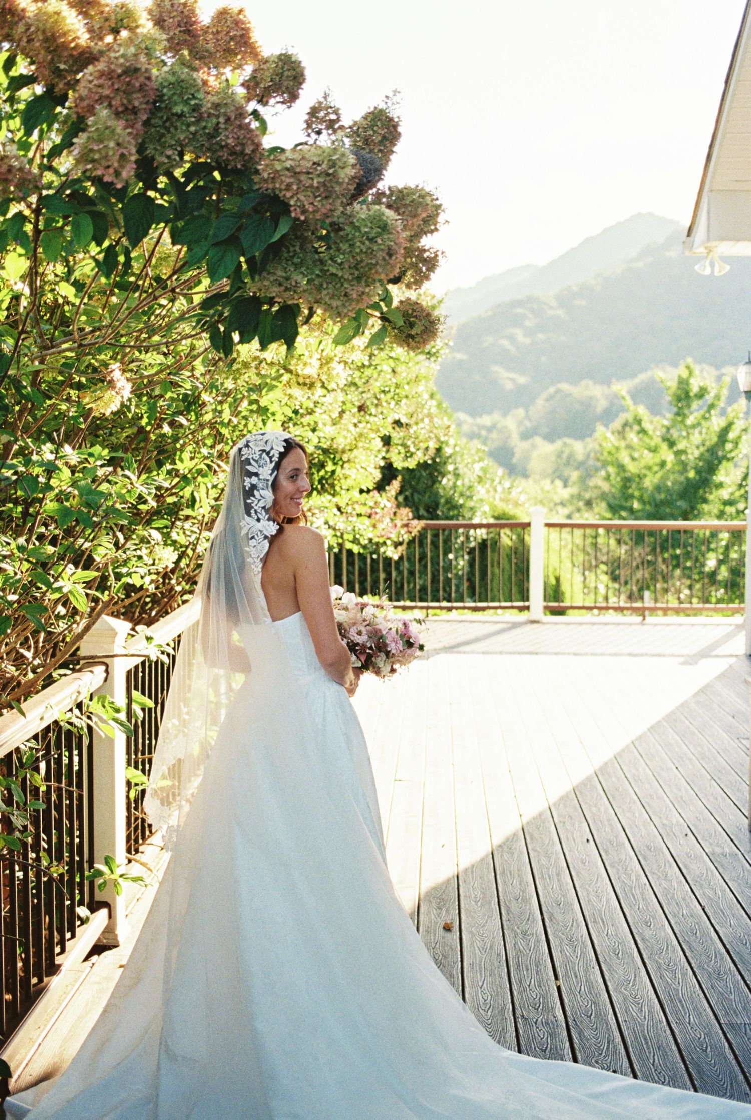 Bride in flowing white gown poses on deck overlooking mountain scenery.