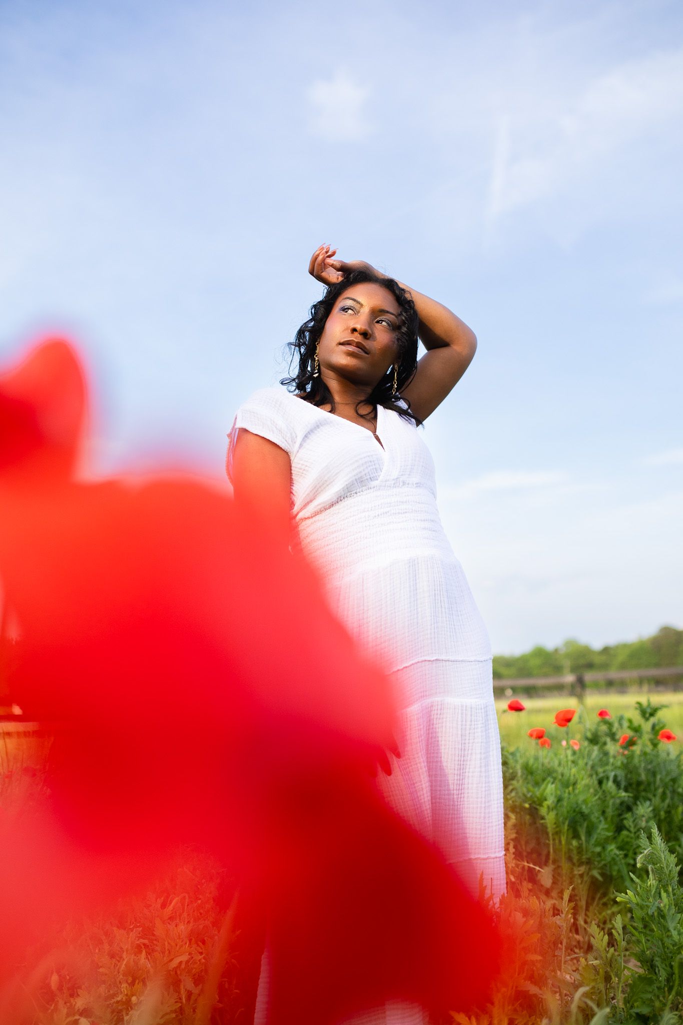 A women in a flowing white dress poses thoughtfully among red poppies at a farm against a blue summer sky.