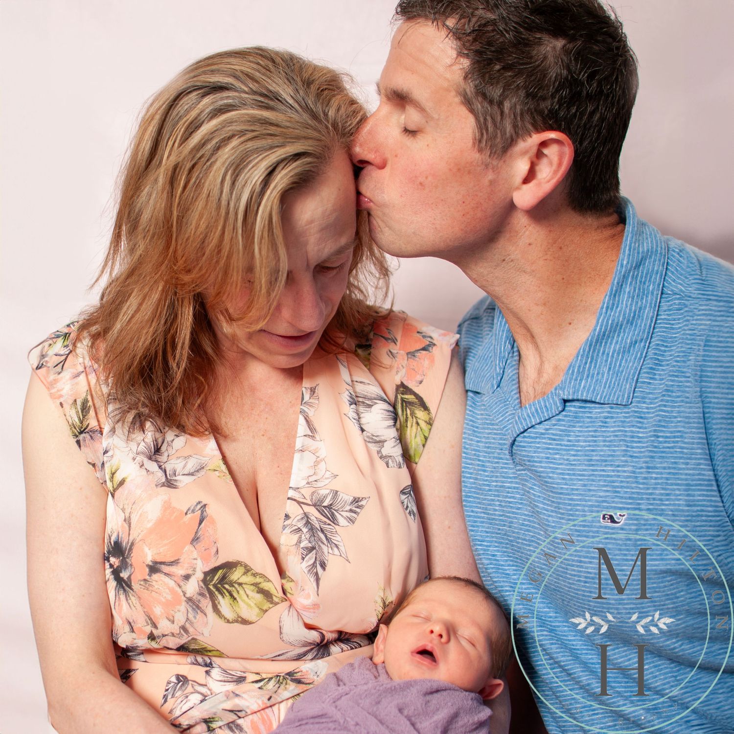 Dad kissing mom's forehead as mom looks at the babyin purple wrap.