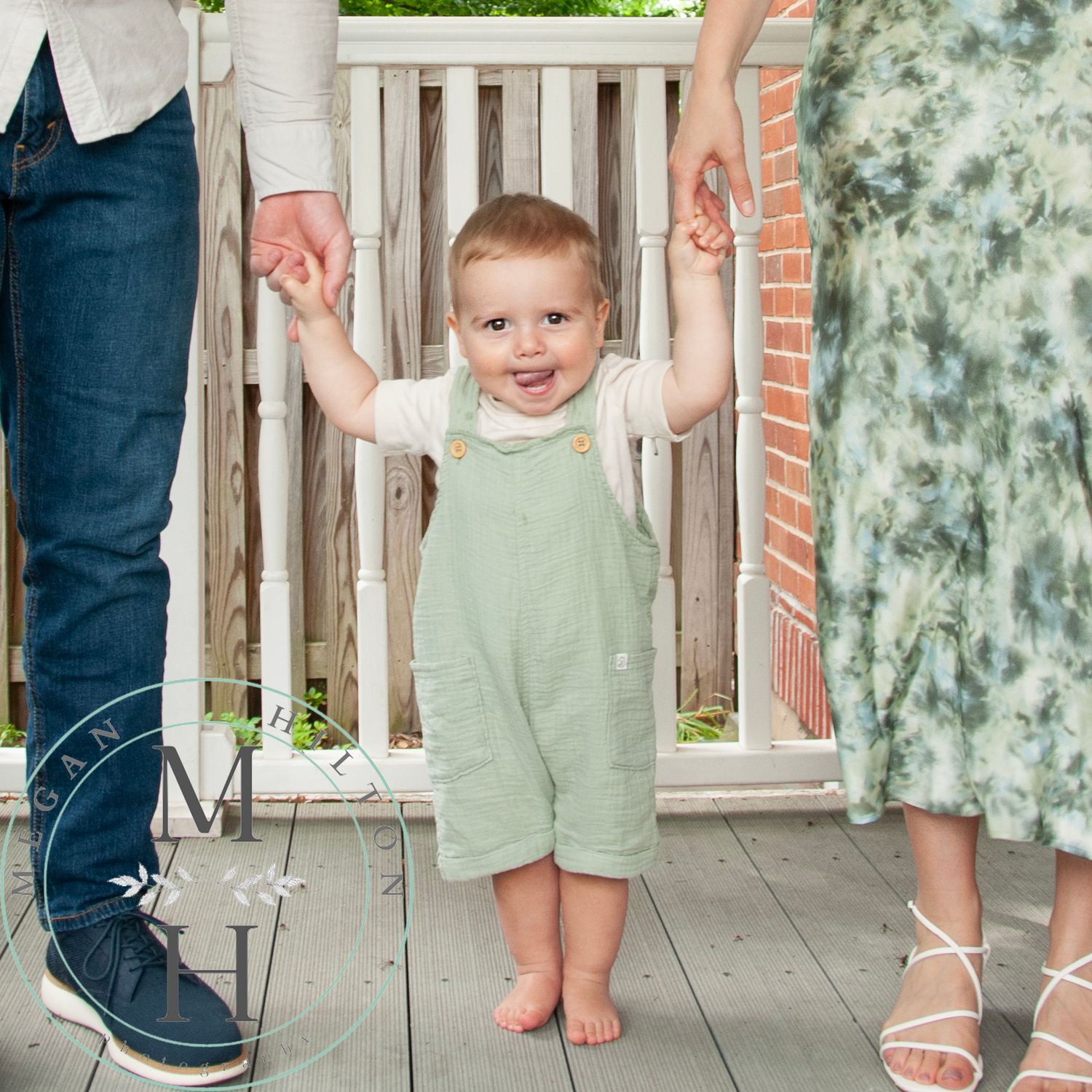 Photo of a smiling toddler in mint green overalls standing barefoot holding parents' hands on a porch with white railing.