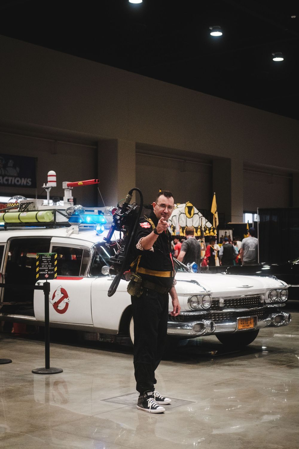 A Ghostbusters cosplayer poses enthusiastically in front of the iconic Ecto-1 car at a convention.