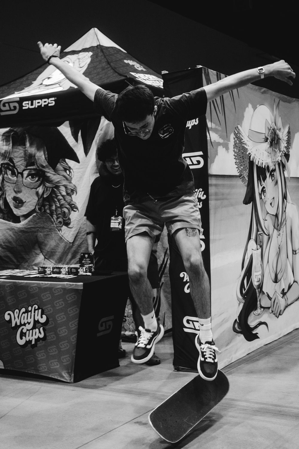 A skateboarder performs a trick against a backdrop with anime-style artwork and graphics at a convention or event.