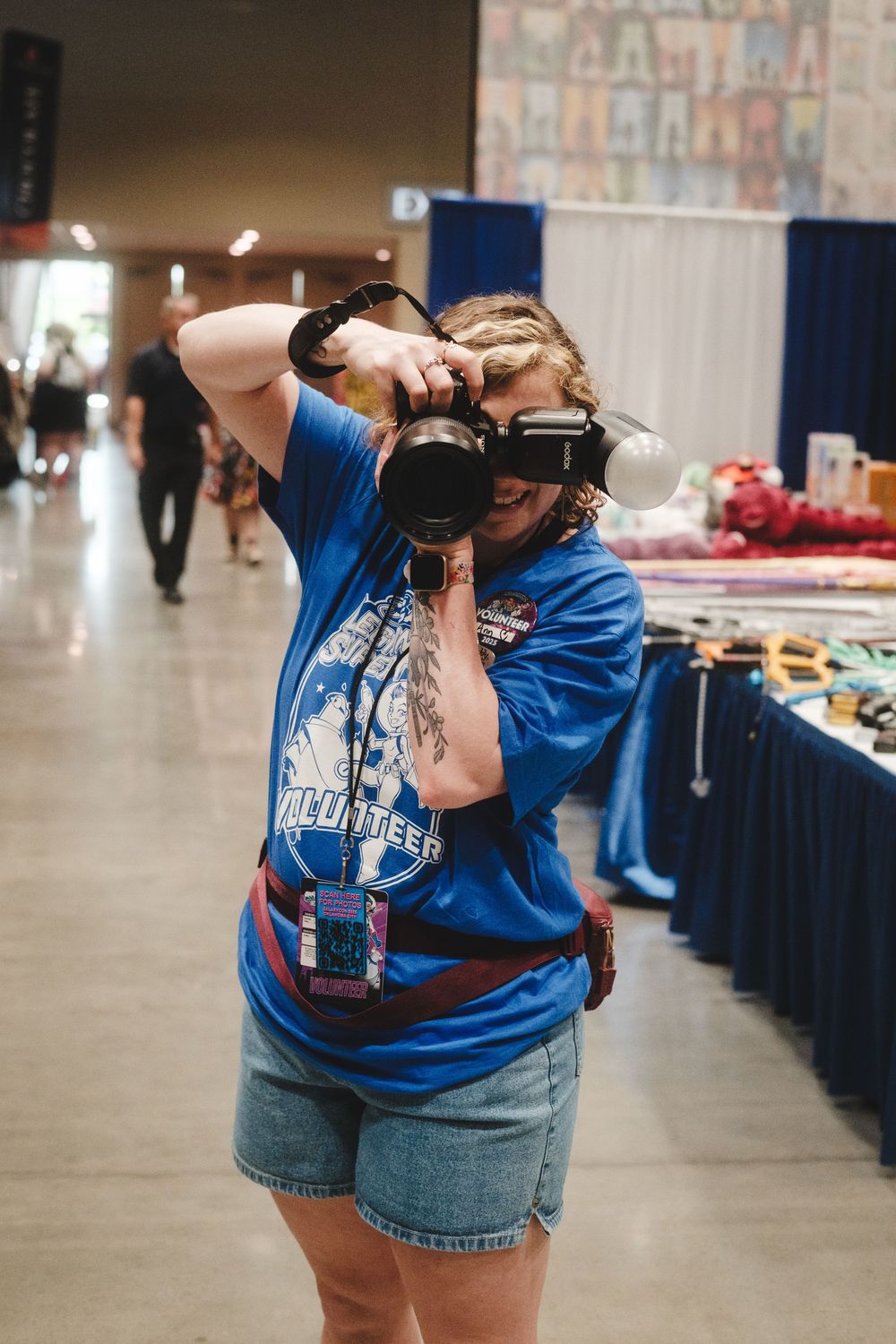 A photographer wearing a blue t-shirt takes photos at a convention with vendor tables visible in the background.