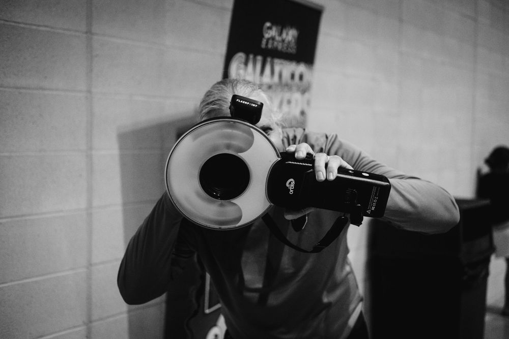 A photographer points a camera with an attached circular flash down a hallway in black and white.