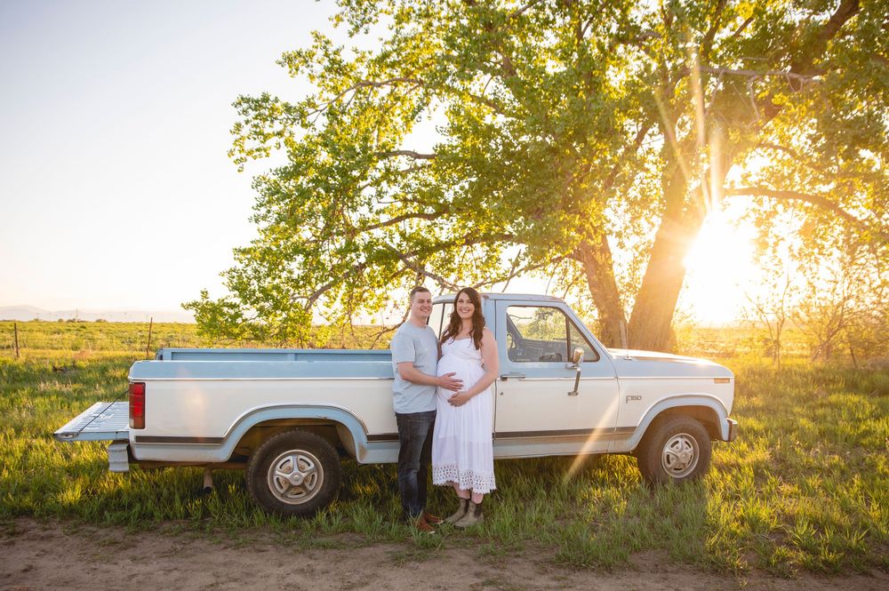 Couple stands beside vintage white pickup truck in golden sunlit field at sunset with large tree in background.