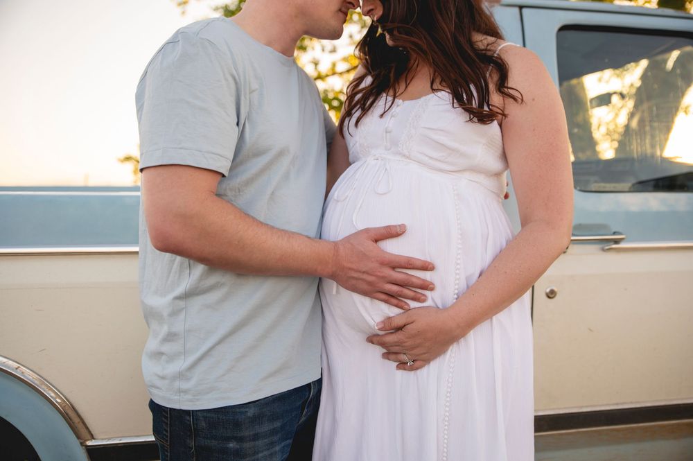 Expectant couple shares tender moment by vintage car as partner gently holds pregnant belly in white dress.