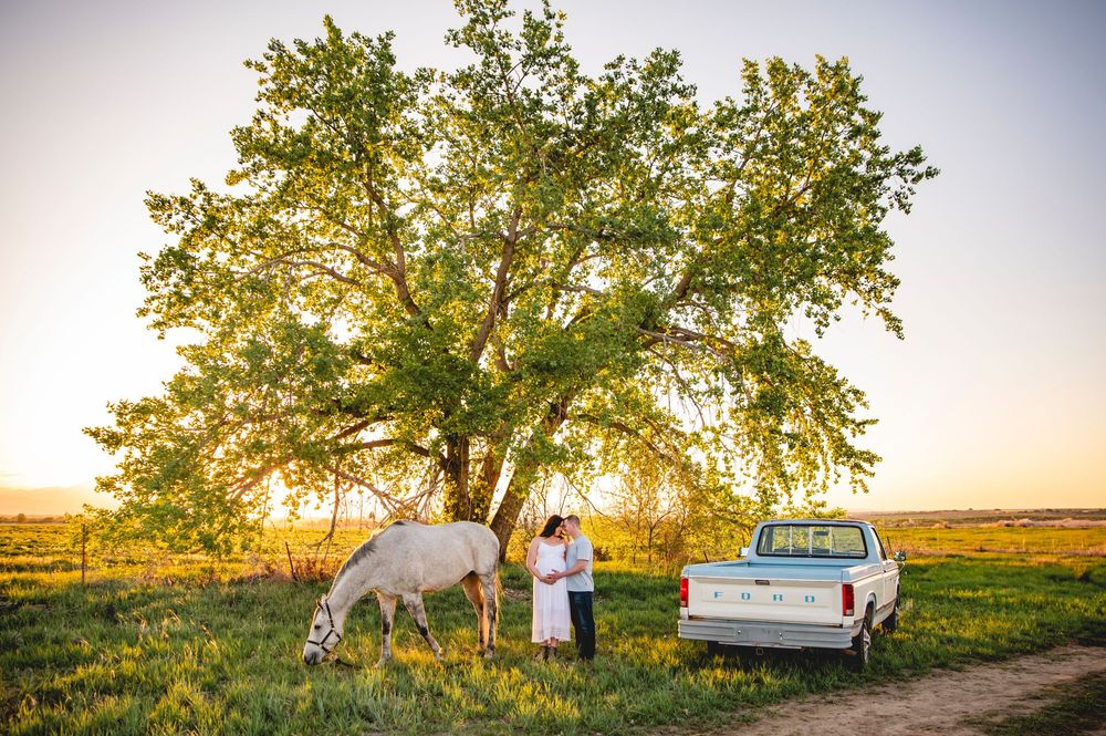 A couple stands with a white horse near a truck and large tree at sunset in a rural field setting.