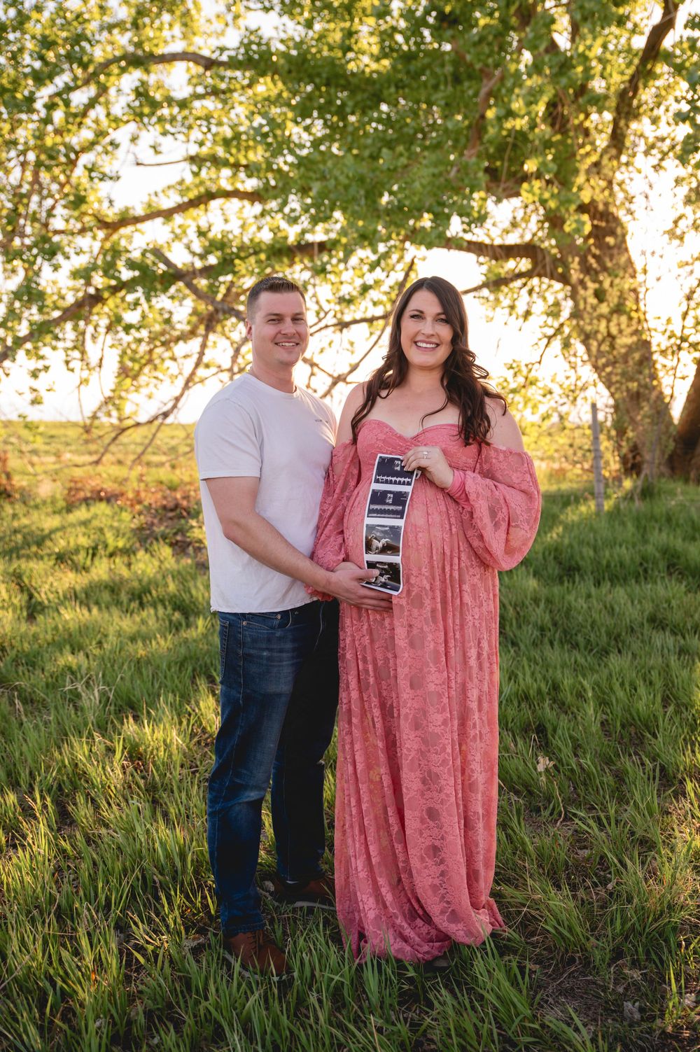 A couple holds an ultrasound photo together in a sunlit field during sunset, sharing a joyful pregnancy announcement moment.