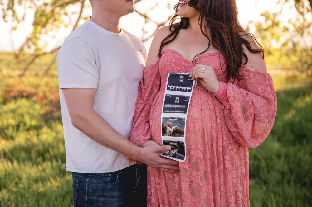 Couple holding ultrasound photos in a sunlit field while wearing pink dress and white t-shirt during sunset.