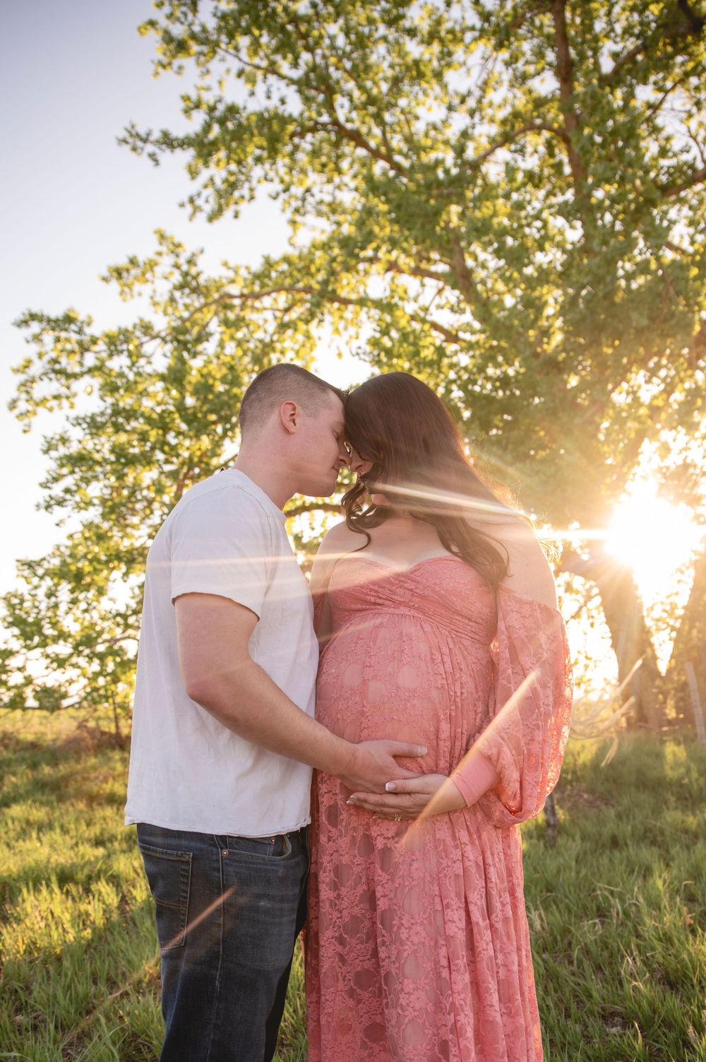 A romantic maternity photo silhouetted against golden sunlight streaming through trees in a grassy field.