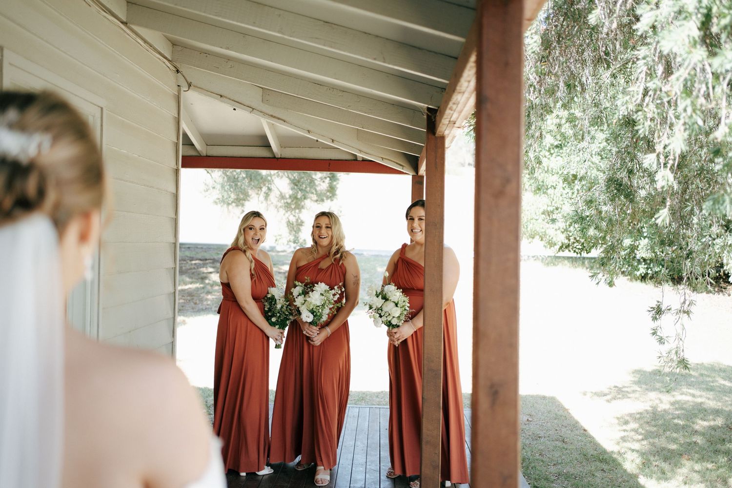 Bridesmaids in rust-colored dresses gather on a covered porch.