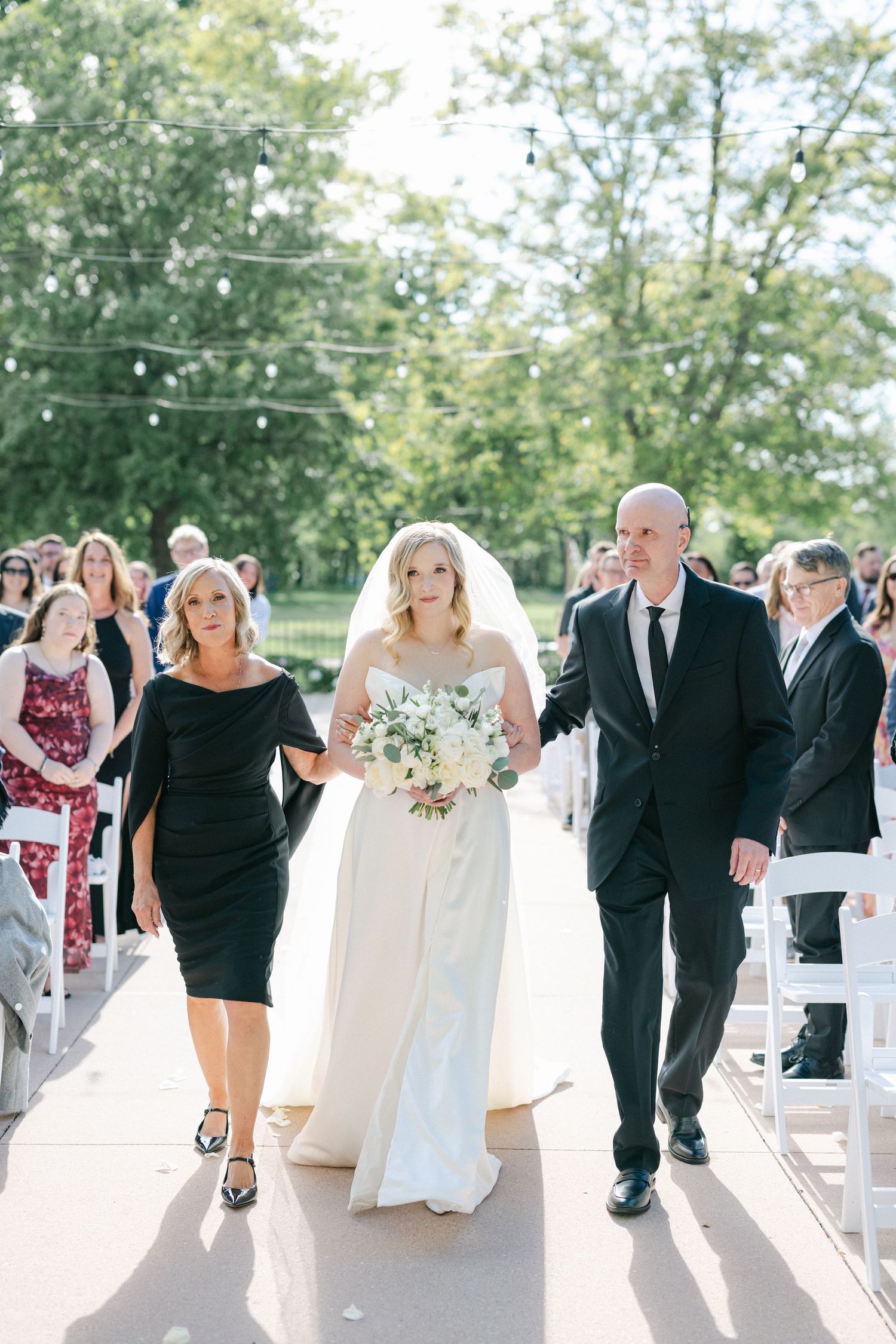 A wedding ceremony on a bright sunny day as people walk down an outdoor aisle lined with white chairs.