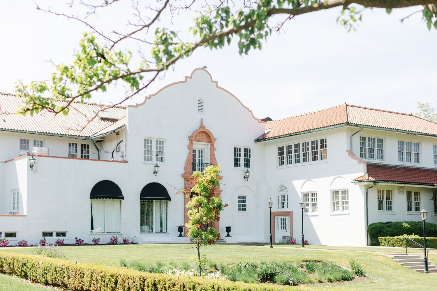 White Spanish colonial style church building with arched entryway and red tile roof nestled among lush greenery. - Ravisloe Country Club