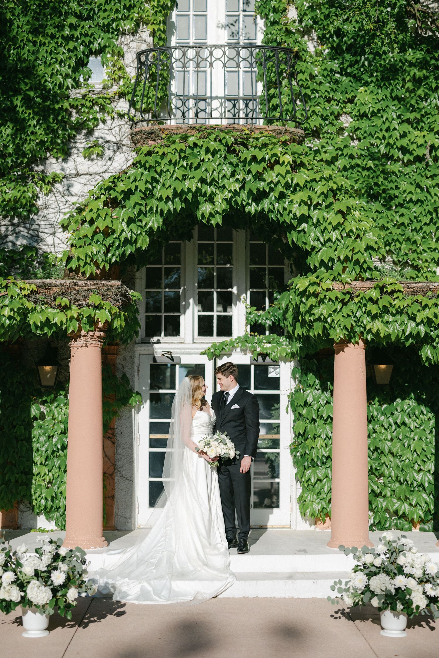 Elegant white wedding dress hangs by window with delicate tulle veil and white rose bouquet arranged below.