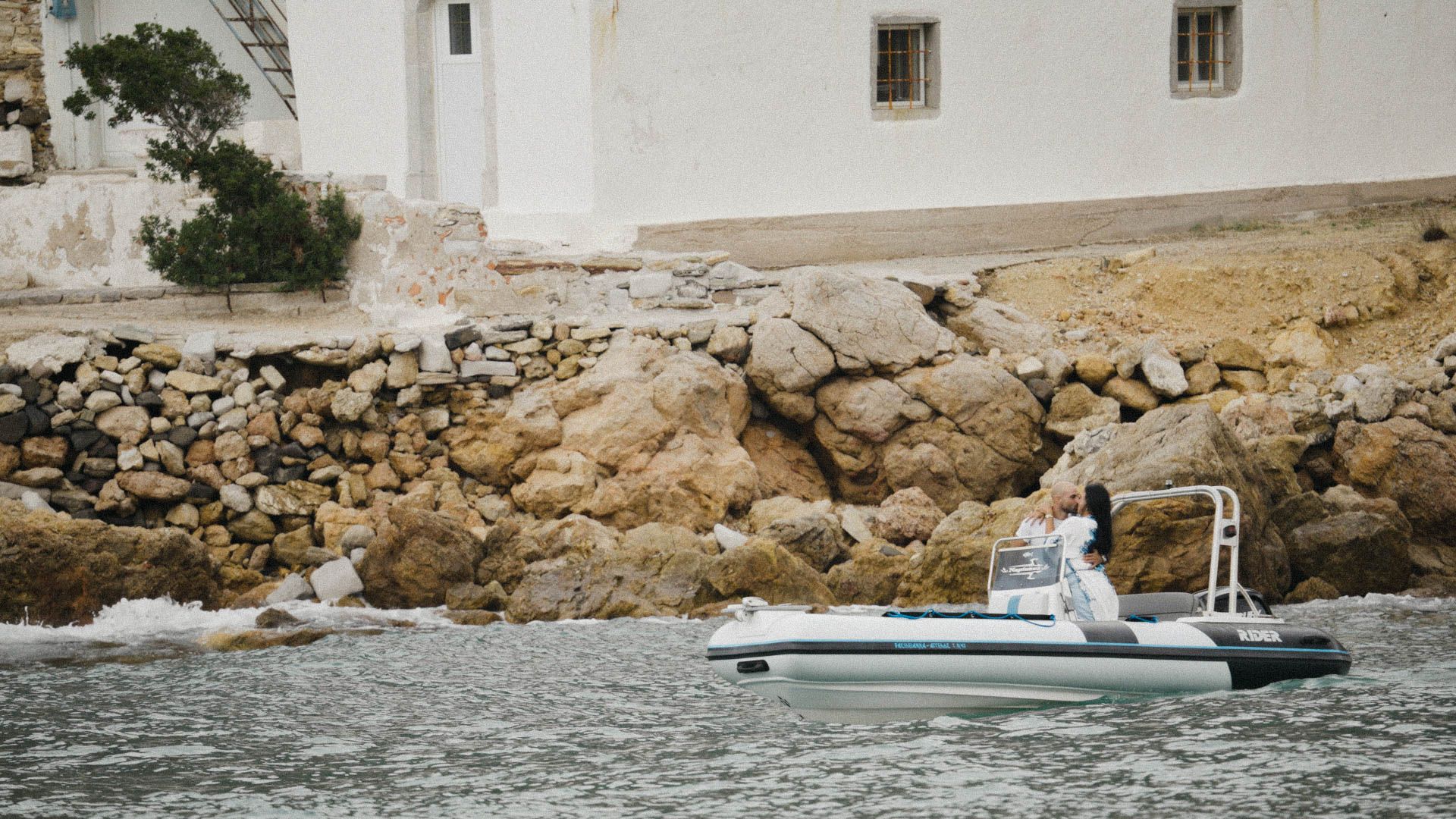 A white boat cruises past rugged coastal rocks and a white building along a Mediterranean shoreline.