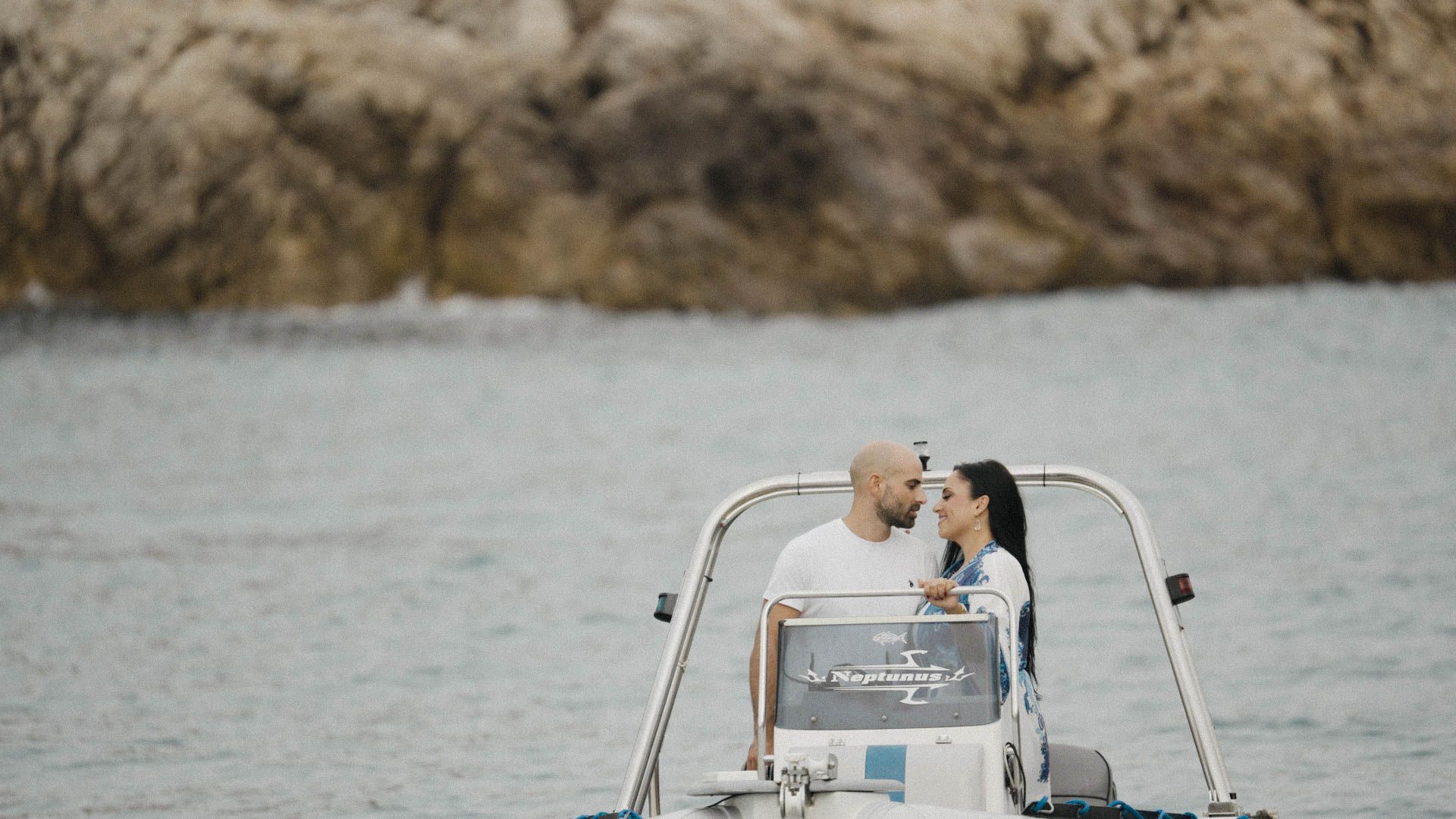 A newly married couple shares a romantic kiss on the deck of a boat with rocky cliffs in the background.