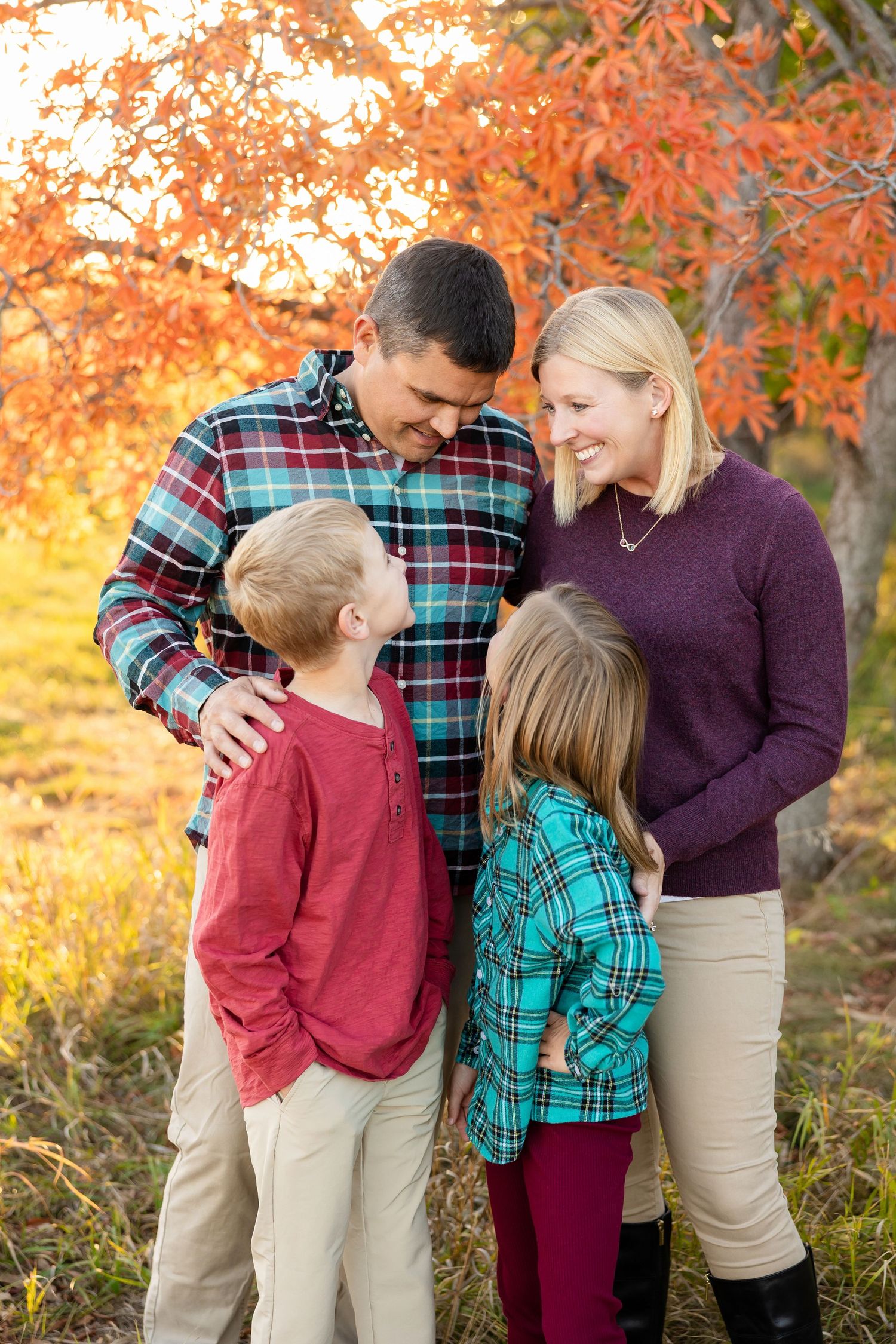 A family of four poses together outdoors during autumn with colorful fall foliage in the background at sunset.