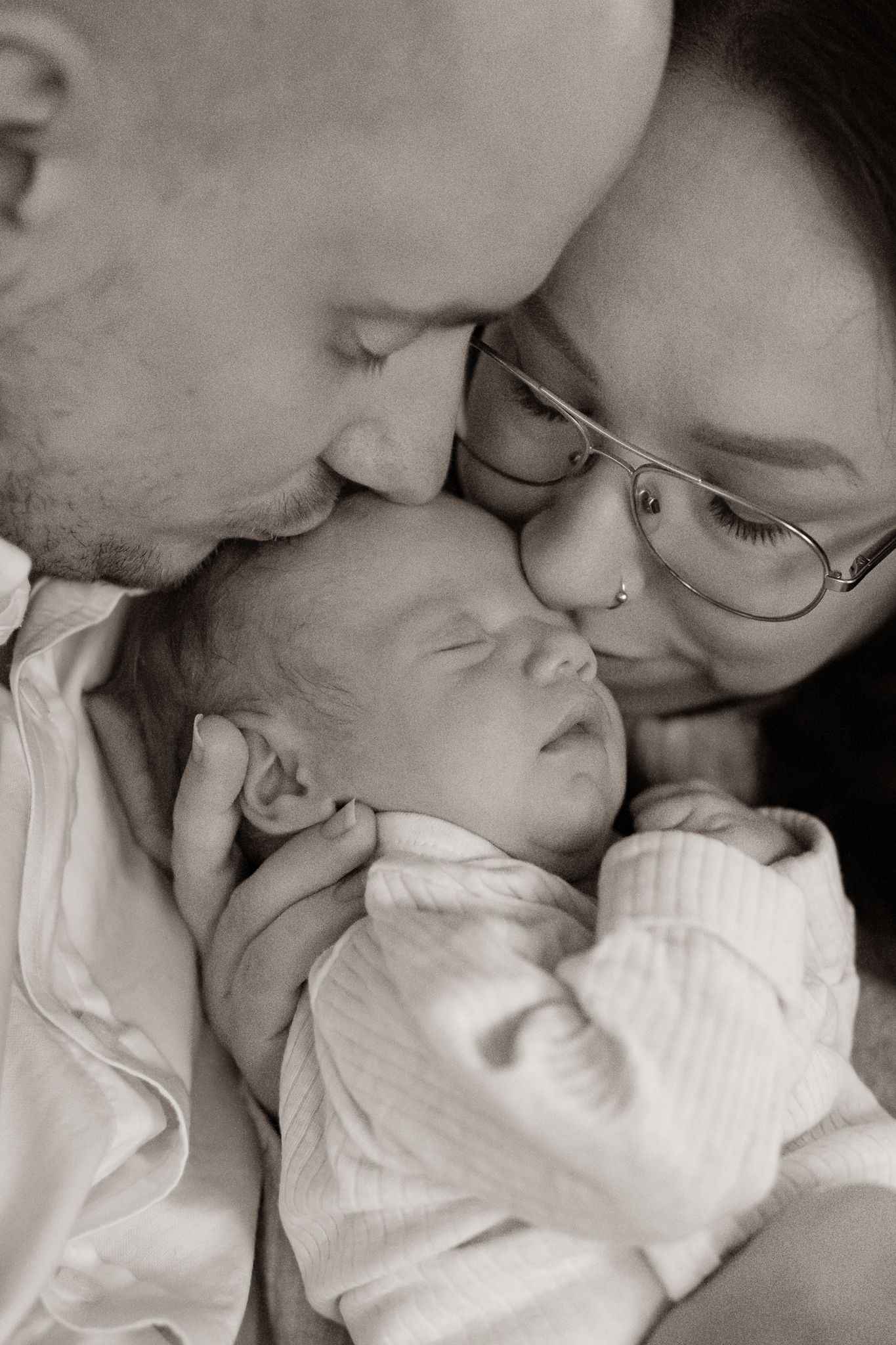 Svartvitt kärleksfullt ögonblick mellan föräldrar och nyfödd sovande bebis, där alla är nära och bebis får pussar på huvudet. Nyföddfotograf