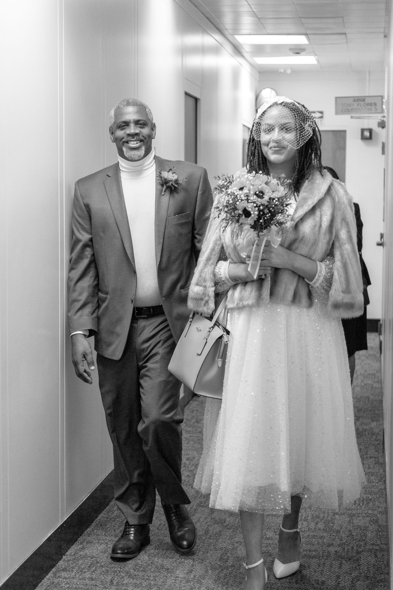 A black and white photo showing two people walking down a hallway, one in a suit and one in a wedding dress with flowers.