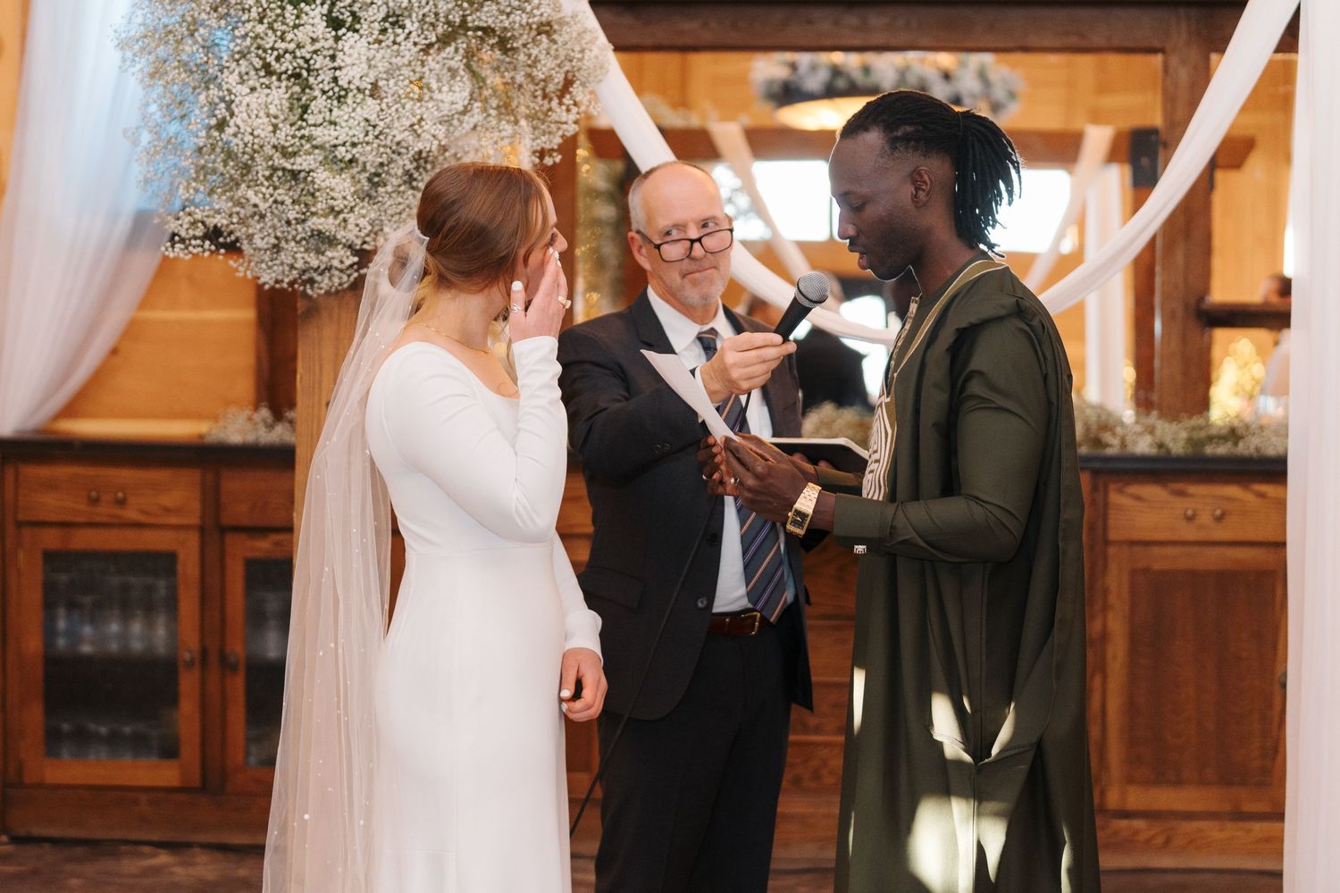 A couple exchanges vows during a wedding ceremony with an officiant in a rustic wooden venue with white decor.