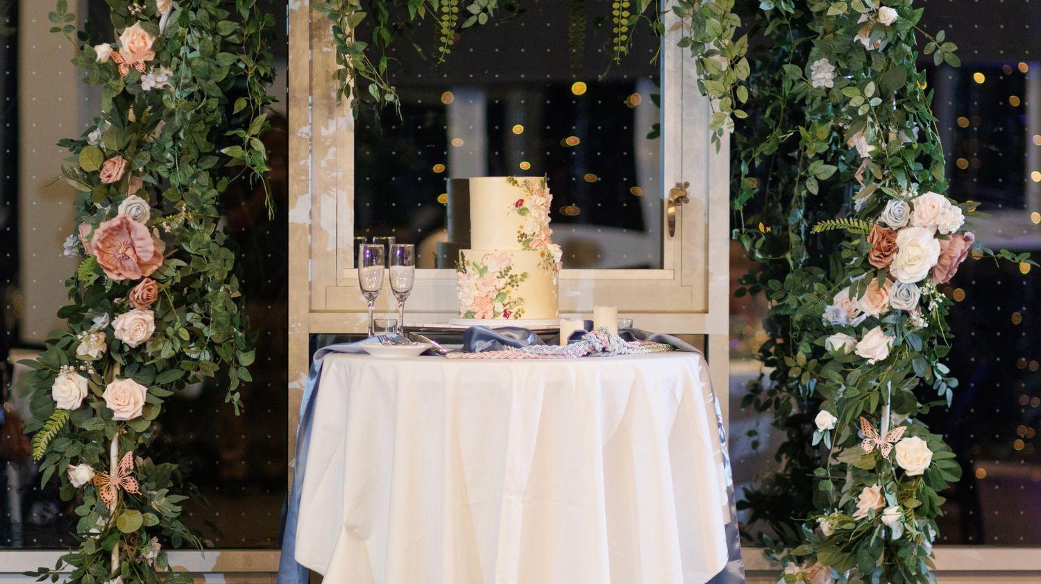 Elegant wedding cake display with floral arrangements and string lights on white tablecloth for outdoor reception.
