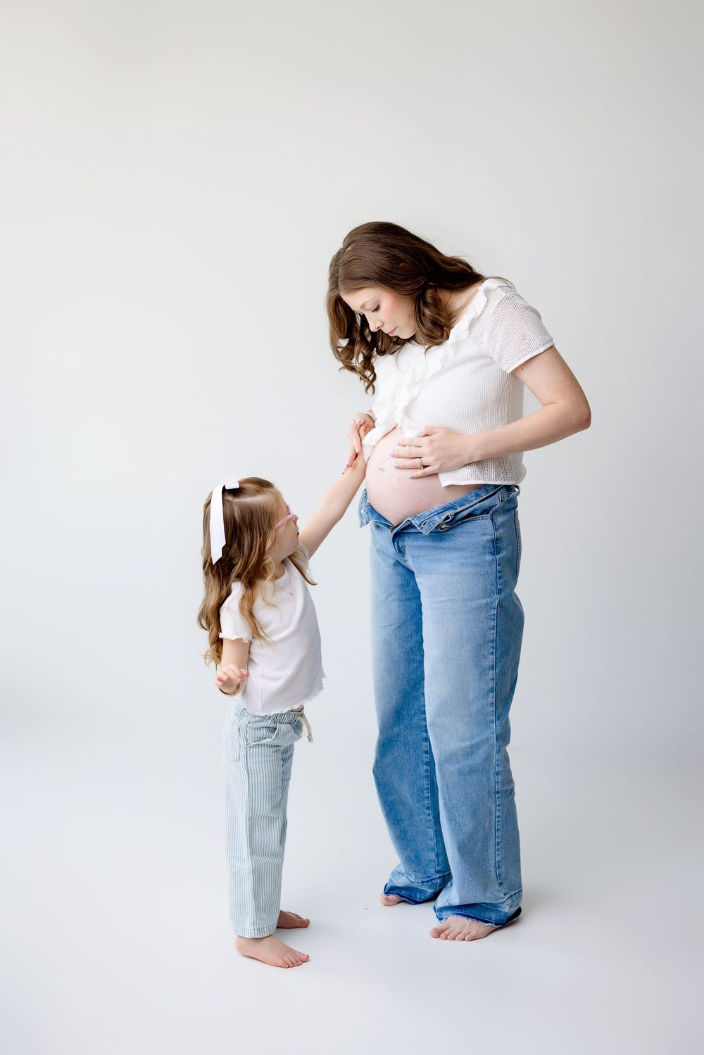 A pregnant woman in jeans and white shirt touches her belly while a young girl in denim reaches up toward her.