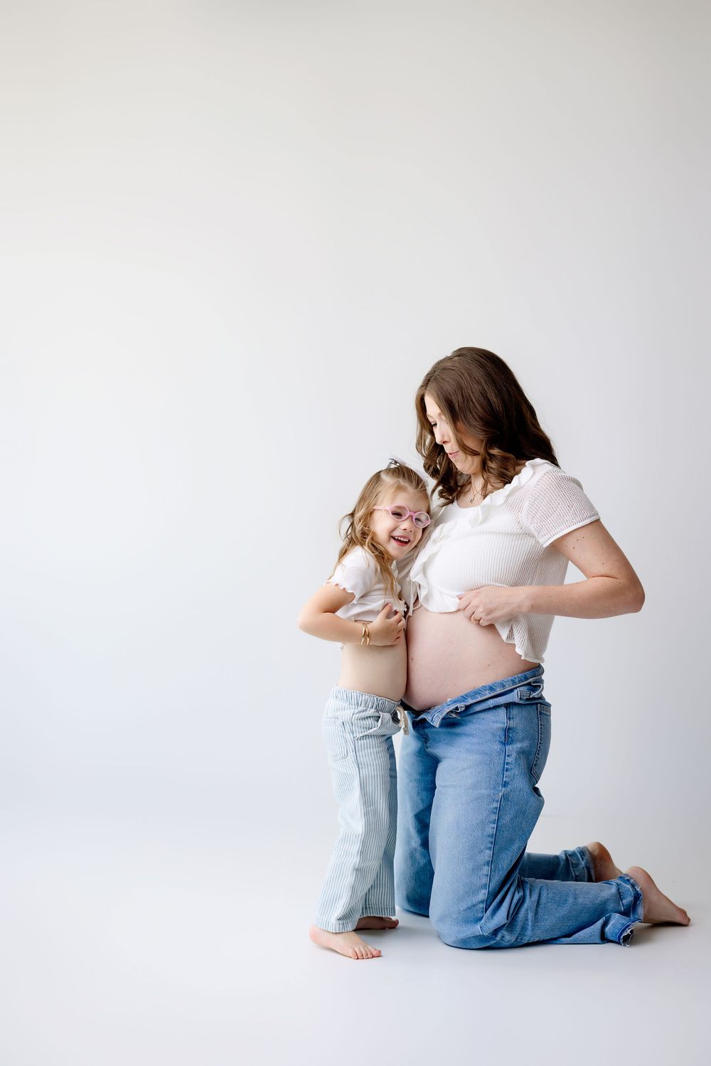 A pregnant woman in jeans and a child in light denim share a joyful moment together against a white backdrop.