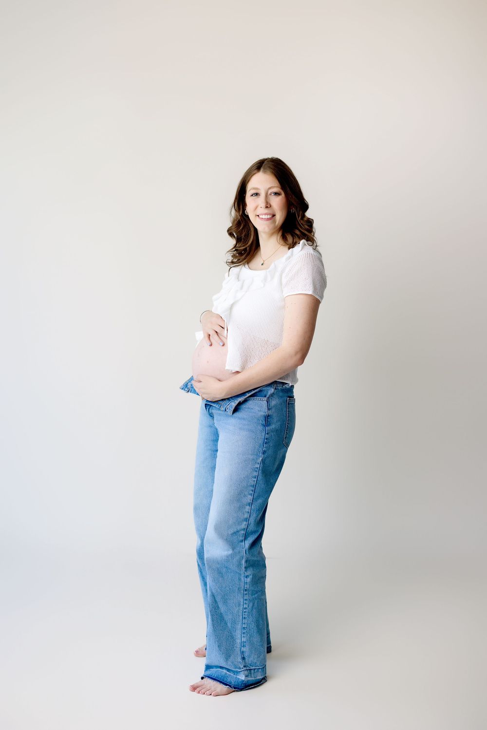 Woman in casual white top and blue jeans poses against light background in simple studio setting.