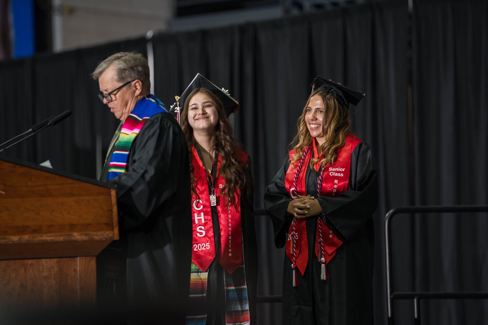 Graduation ceremony showing a speaker at podium with two graduates wearing red stoles on stage at commencement.