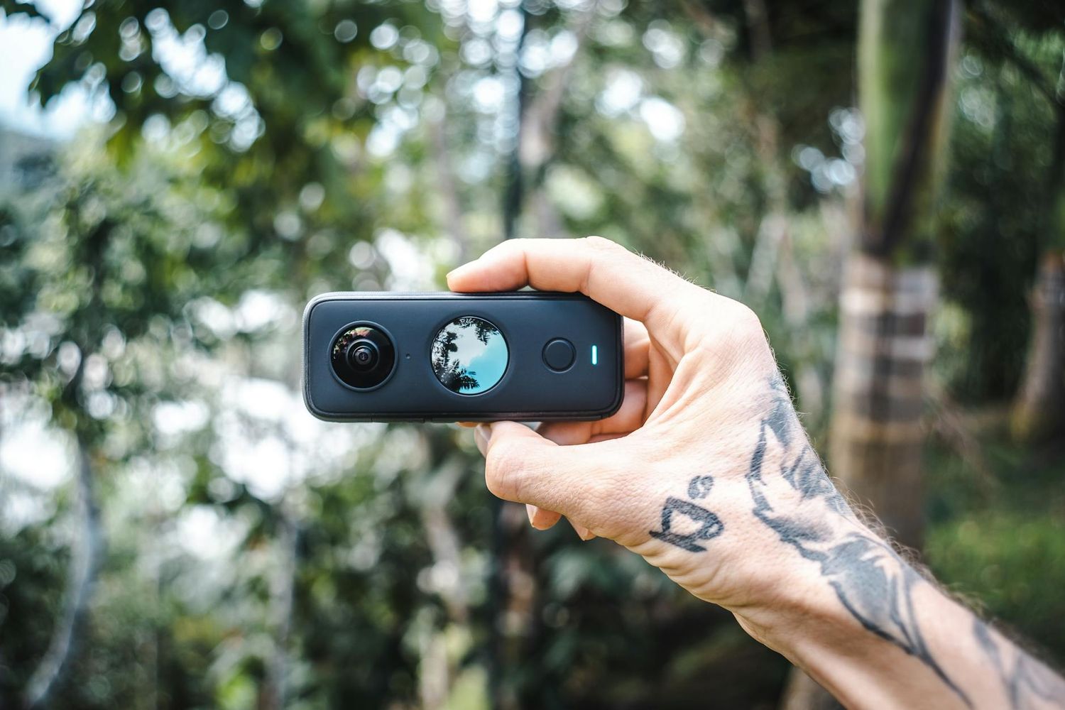 A tattooed hand holds up a small black Insta360 camera outdoors against a blurred green foliage background. Travel Photographer videographer