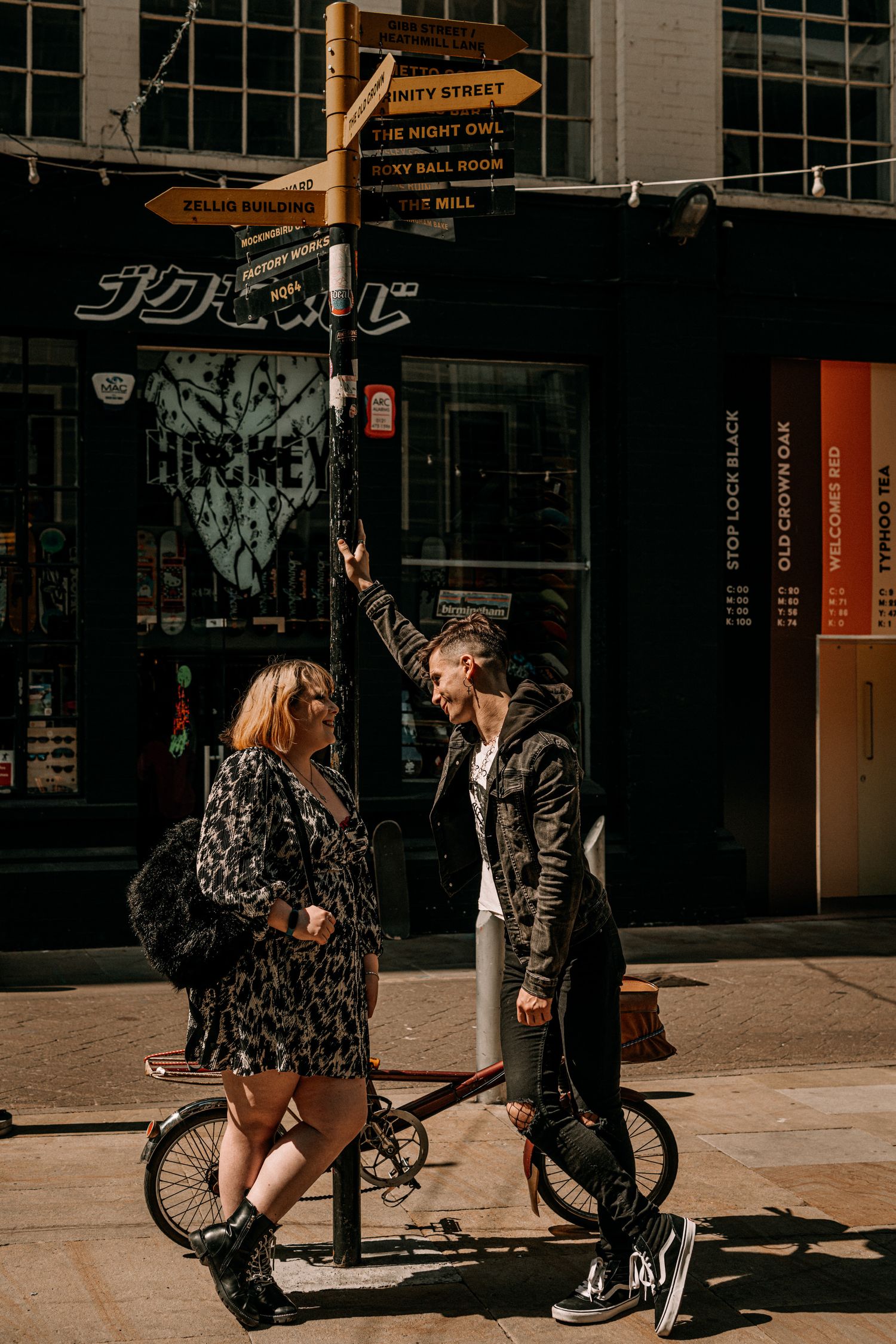 Two people chat while standing near a bicycle on a sunny urban street with storefronts in the background.