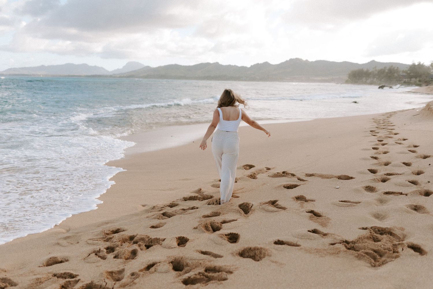 Person in white outfit walking barefoot on beach leaving footprints in sand.